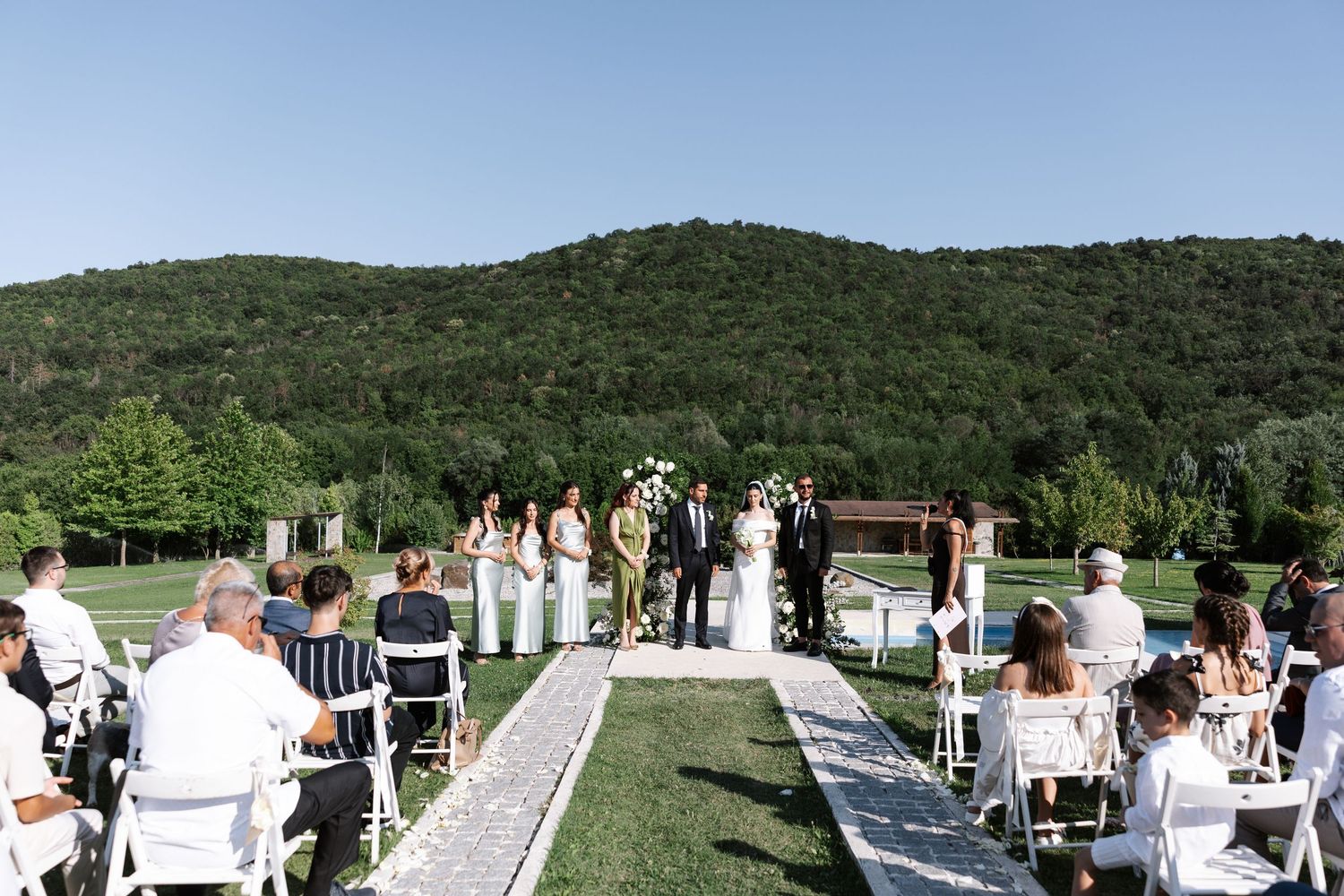 Panoramic view of an outdoor wedding ceremony with guests seated in white chairs against a backdrop of rolling green hills.