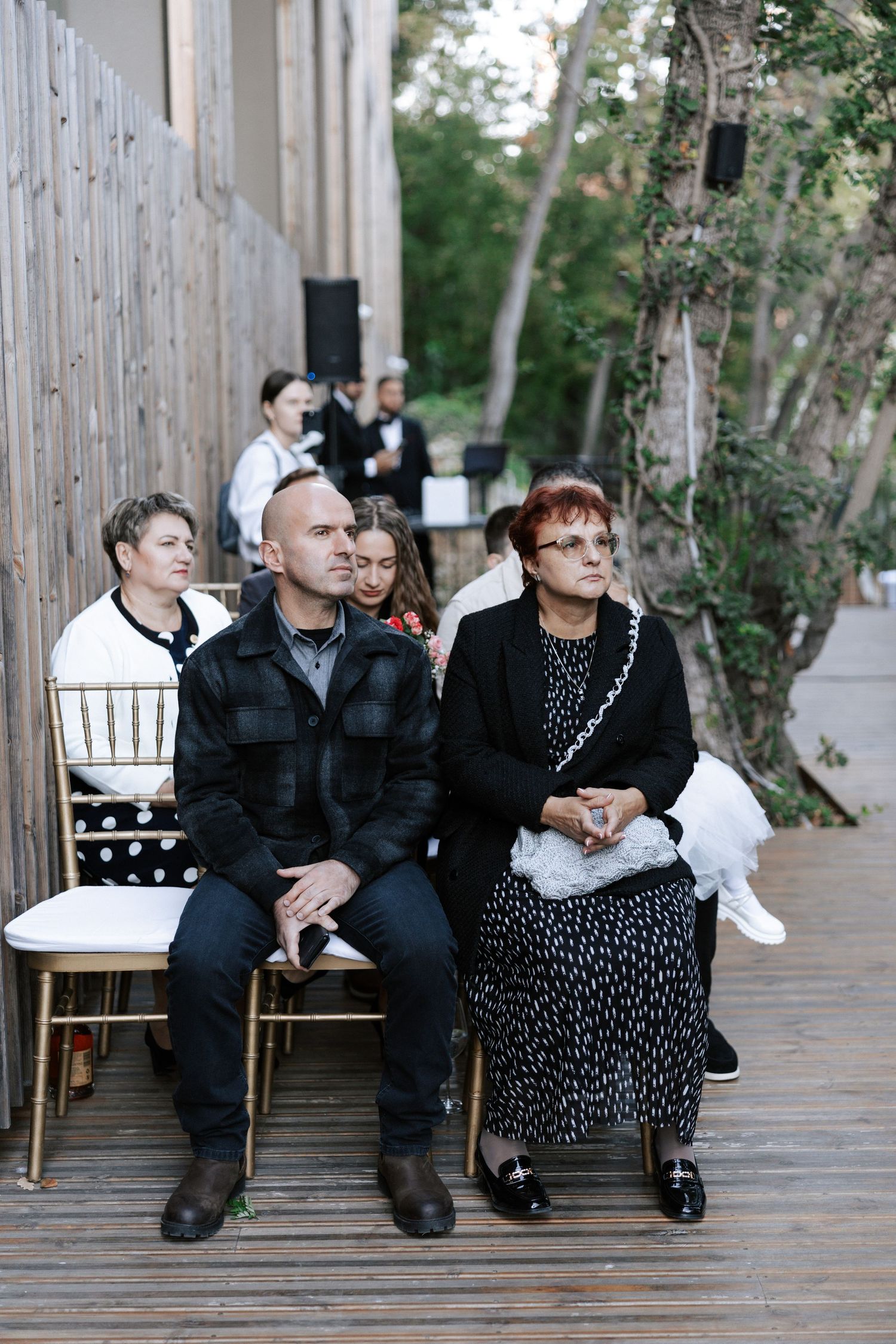 Wedding guests sit on wooden chairs on an outdoor deck during a ceremony.