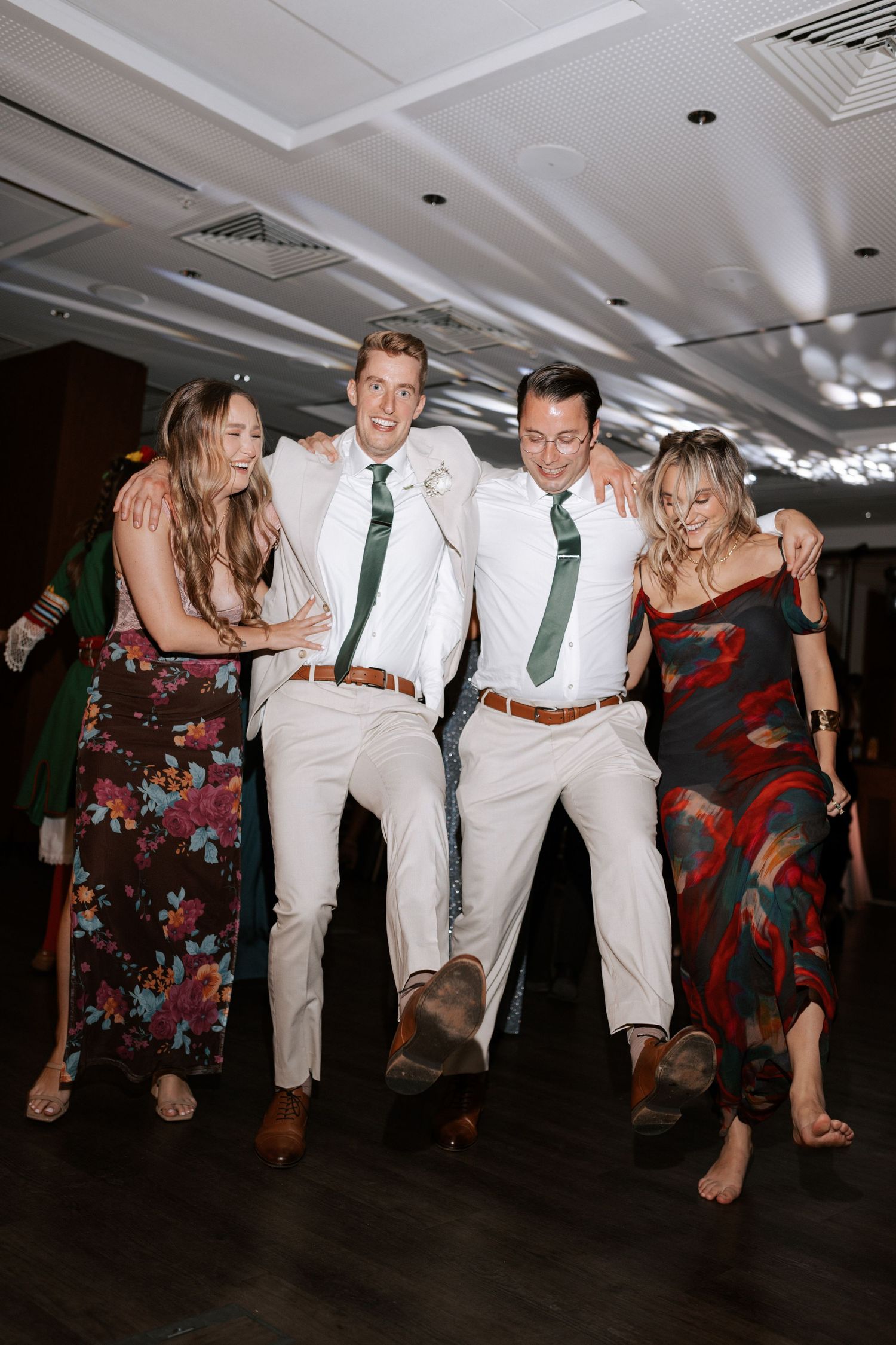 Party guests in formal attire dance energetically on a dark wooden floor at a wedding reception.