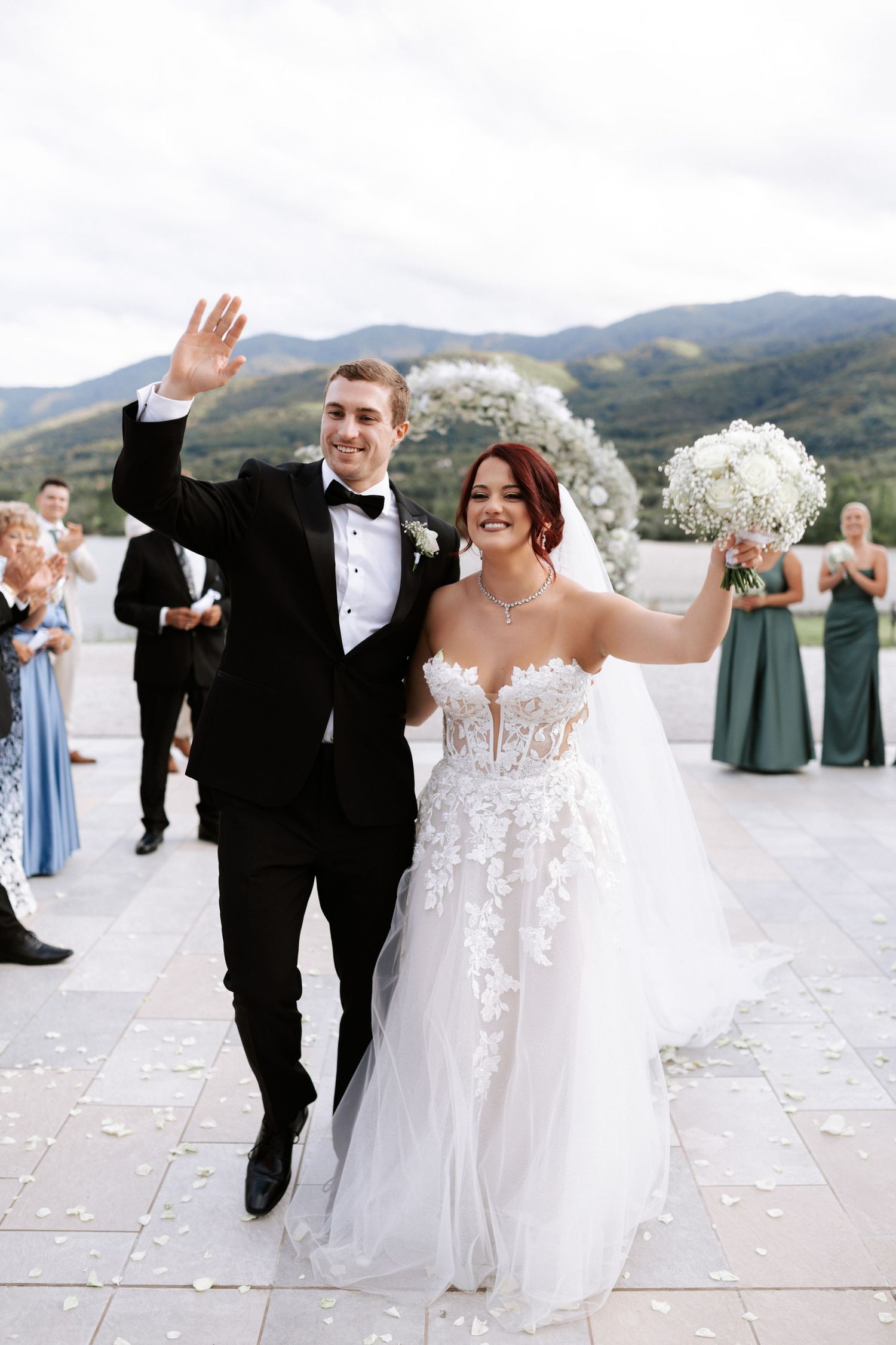 Wedding couple waves to guests during outdoor celebration with mountain backdrop.