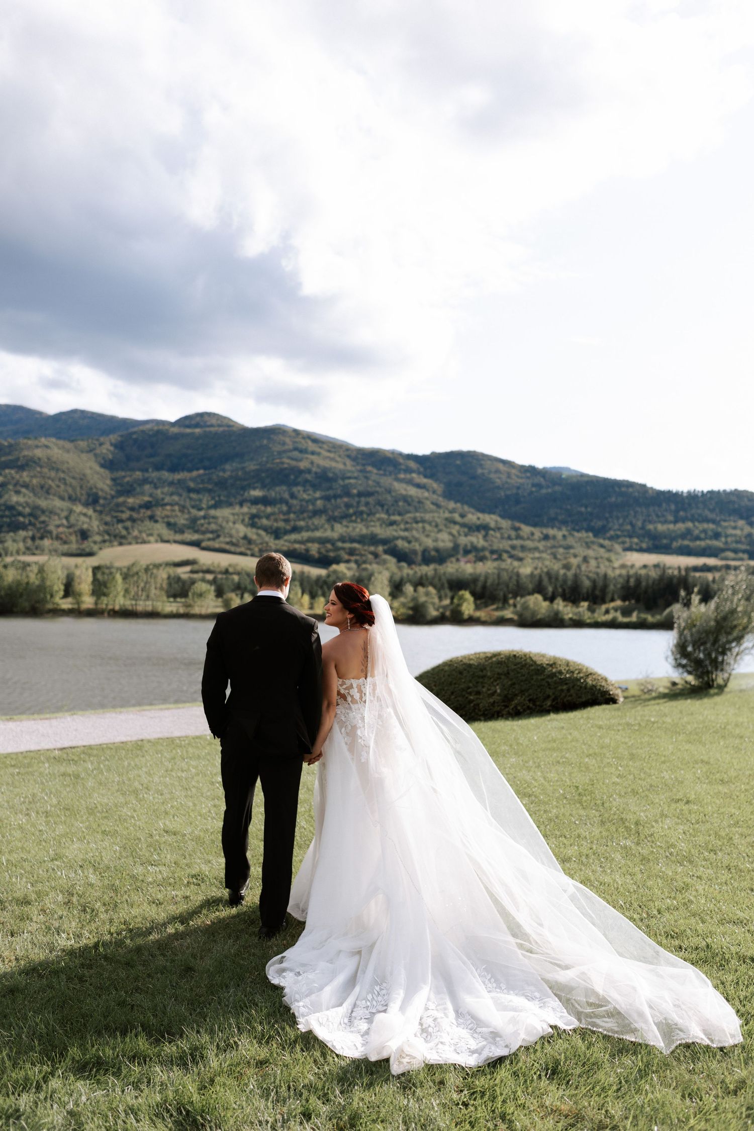 Couple stands overlooking mountain lake landscape in elegant wedding attire.