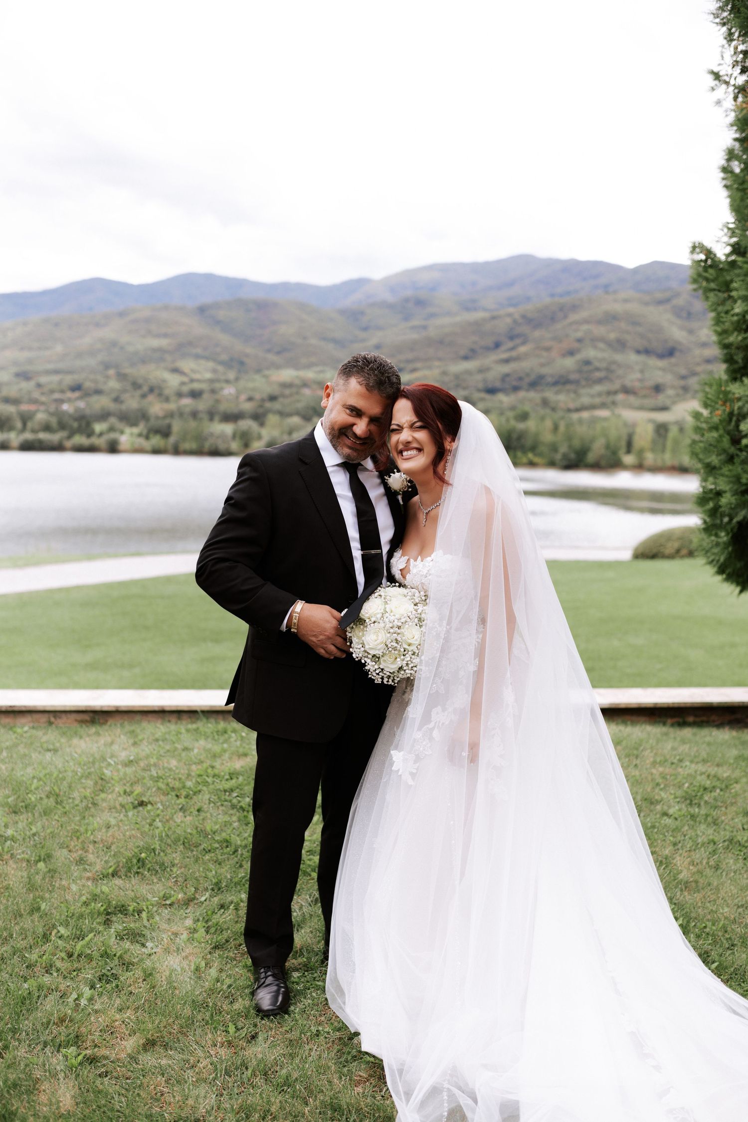 Wedding couple shares romantic moment on lawn with scenic mountain lake backdrop.