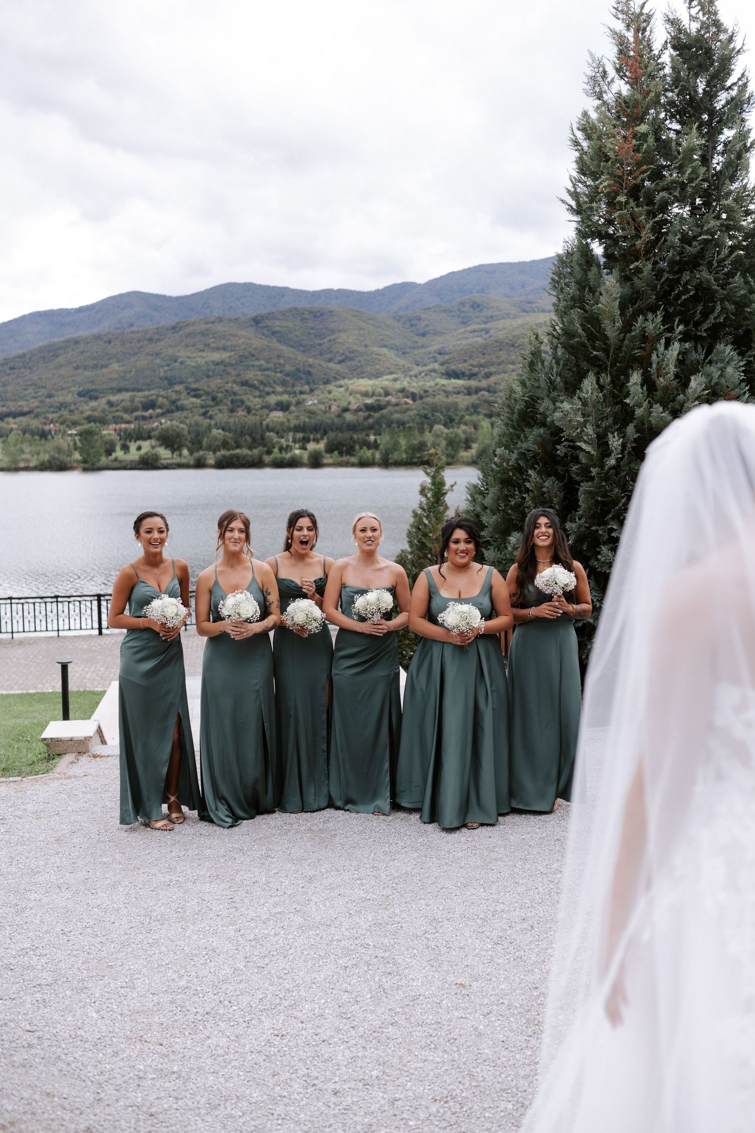 Wedding party in sage green dresses poses by a lakeside mountain venue.