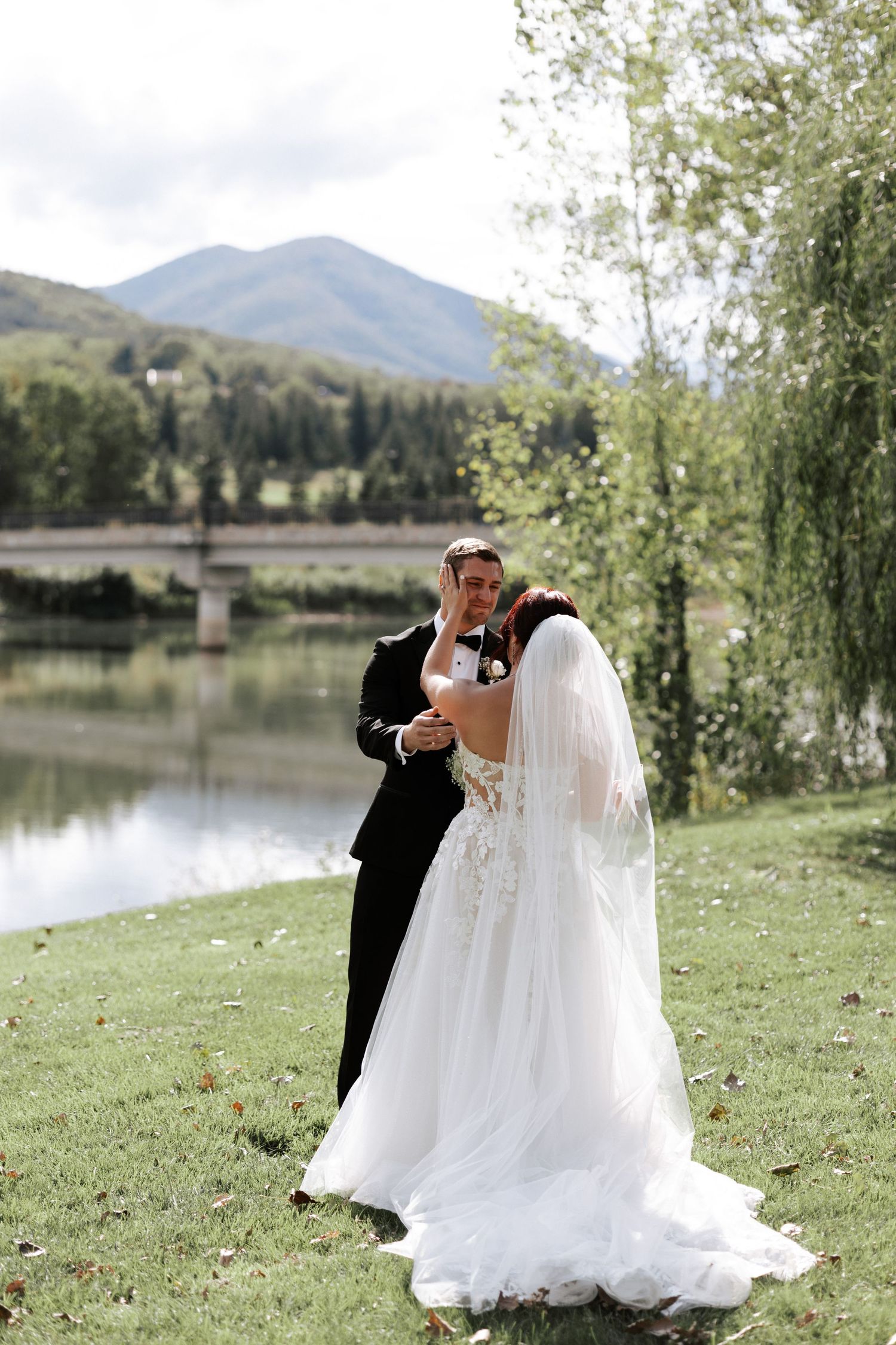 A newlywed couple embraces on a lush lawn with a scenic lake and mountain vista.