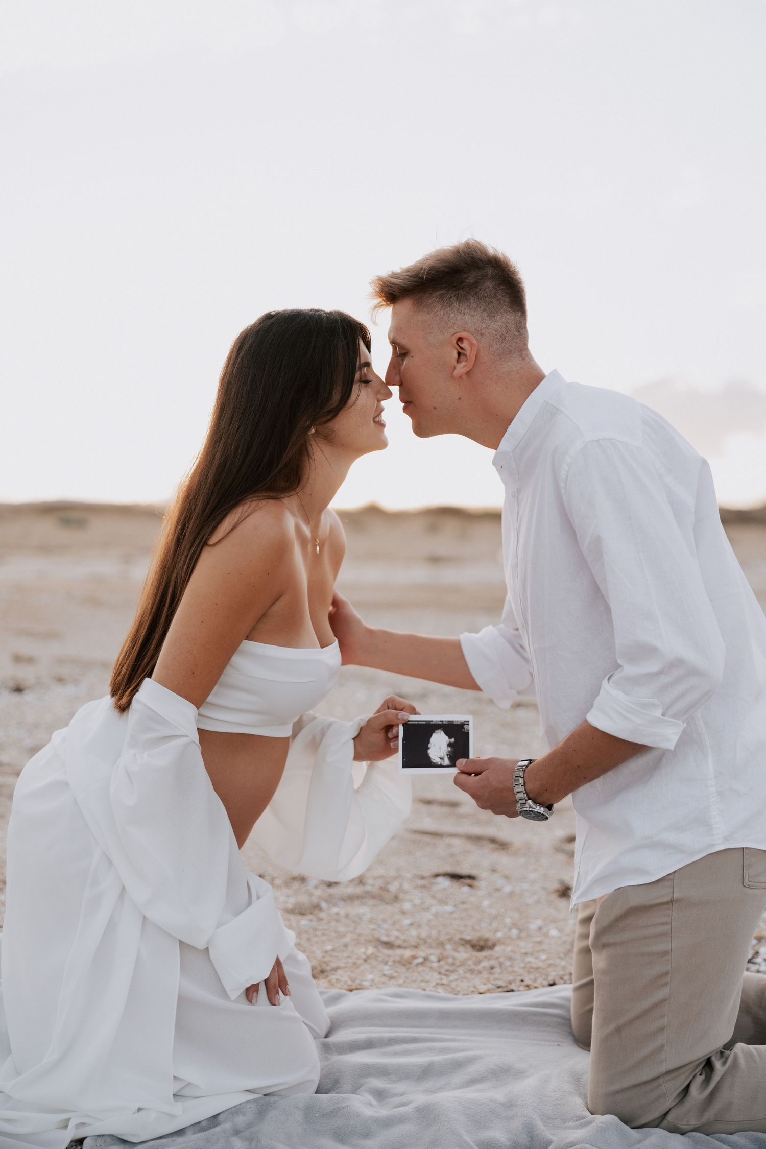 An expecting couple shares a moment on the beach while holding an ultrasound photo.