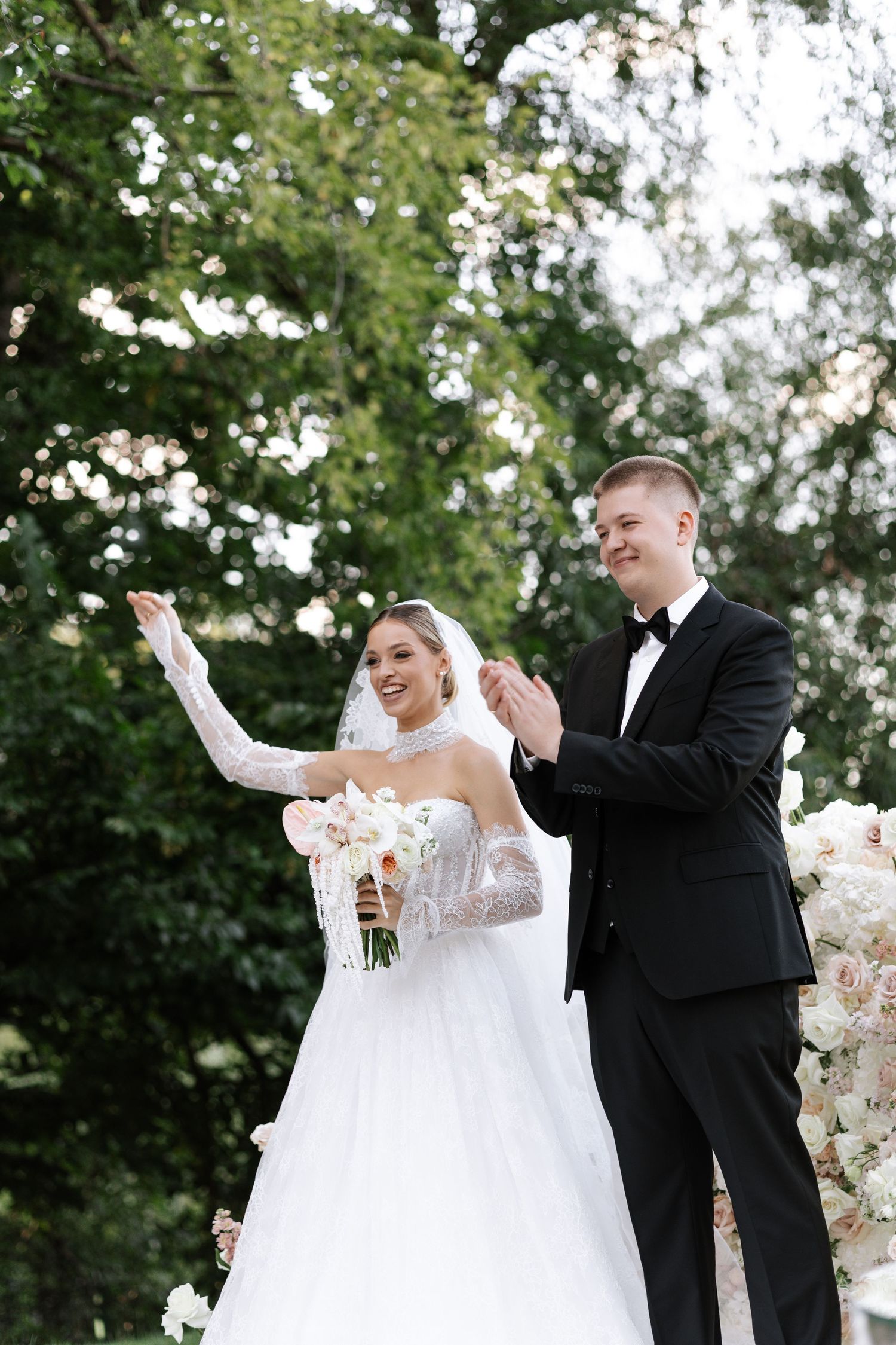 A joyful wedding couple shares a candid moment outdoors surrounded by greenery and white floral arrangements.