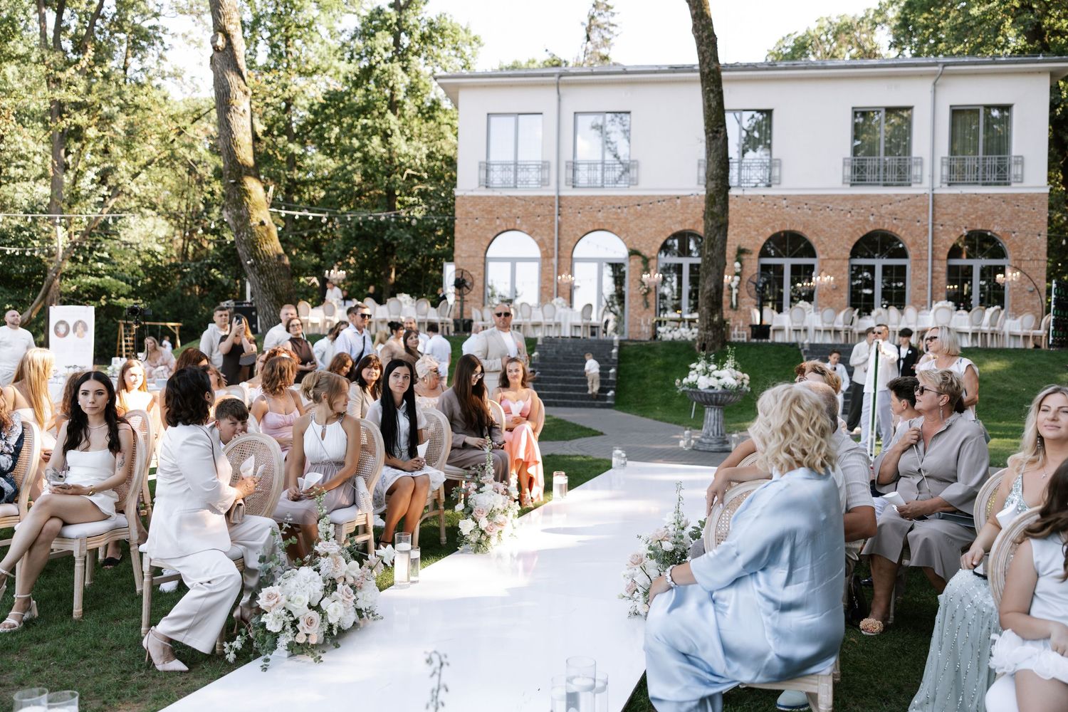 Outdoor wedding ceremony setup with white chairs and guests on a sunny garden lawn.