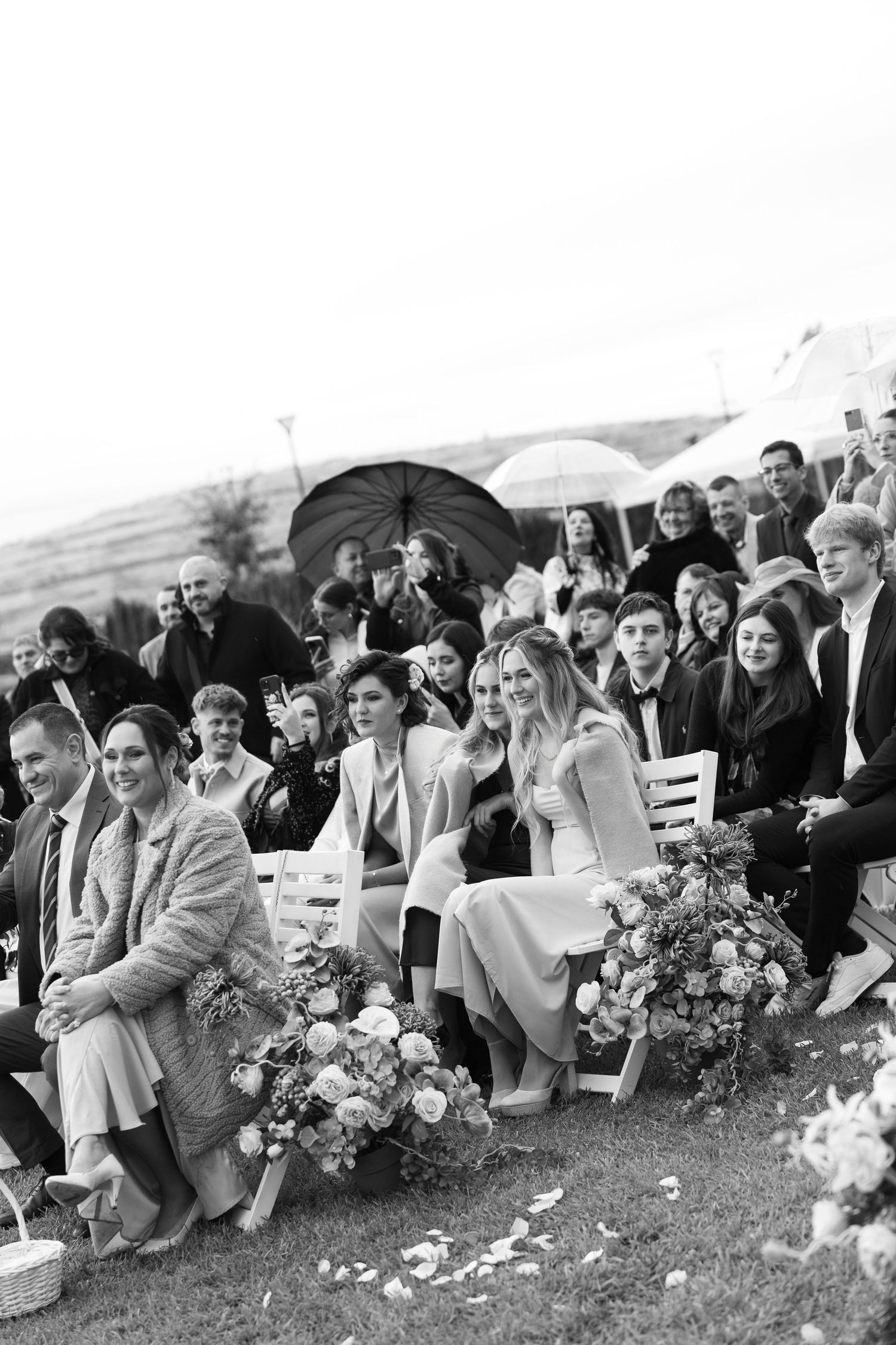 A large group of wedding guests sit together on a grassy hill while holding floral bouquets at an outdoor celebration.