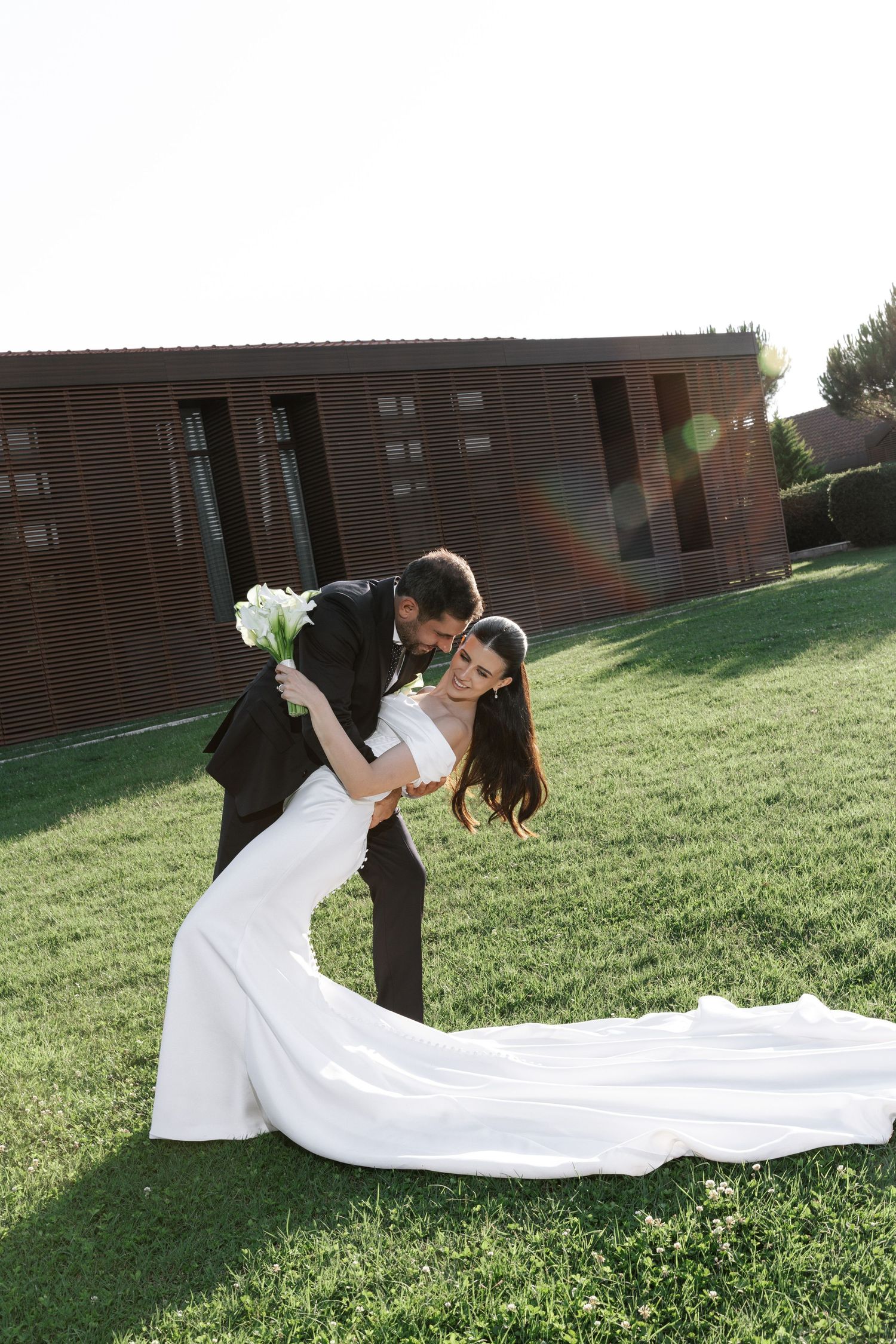 A romantic wedding couple dips and poses on a grassy lawn with a flowing white dress train trailing behind them.