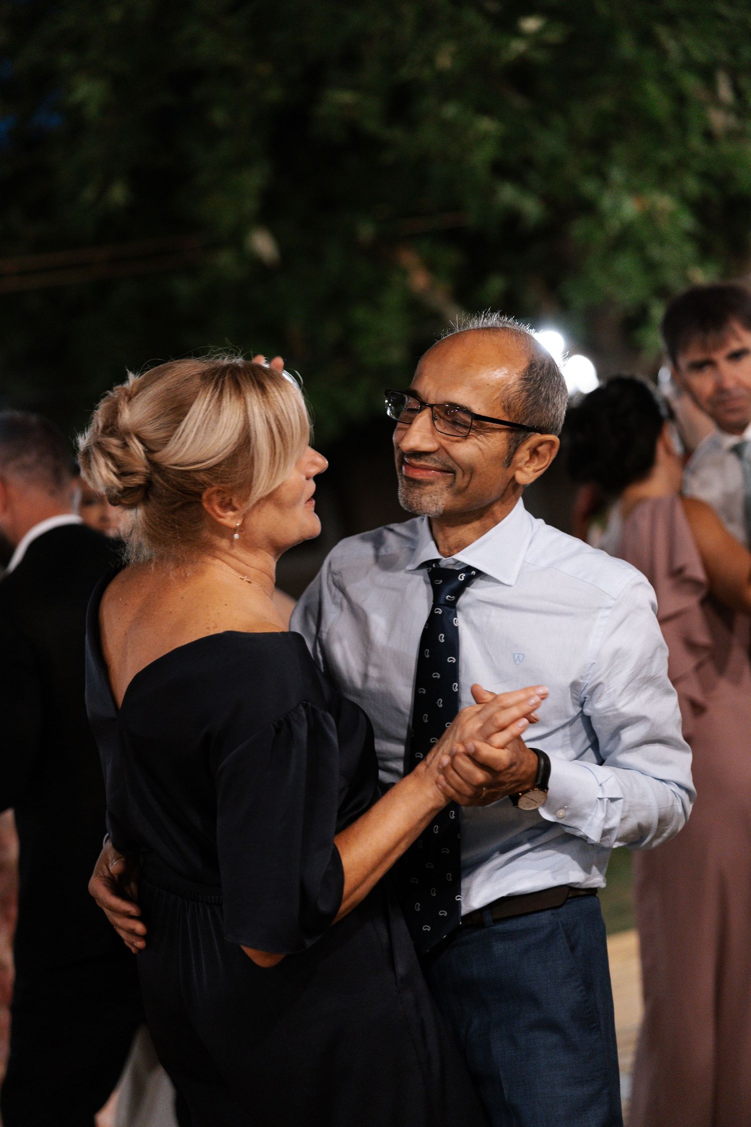 A couple dancing together at an outdoor event, sharing a romantic moment while other guests mingle in the background.
