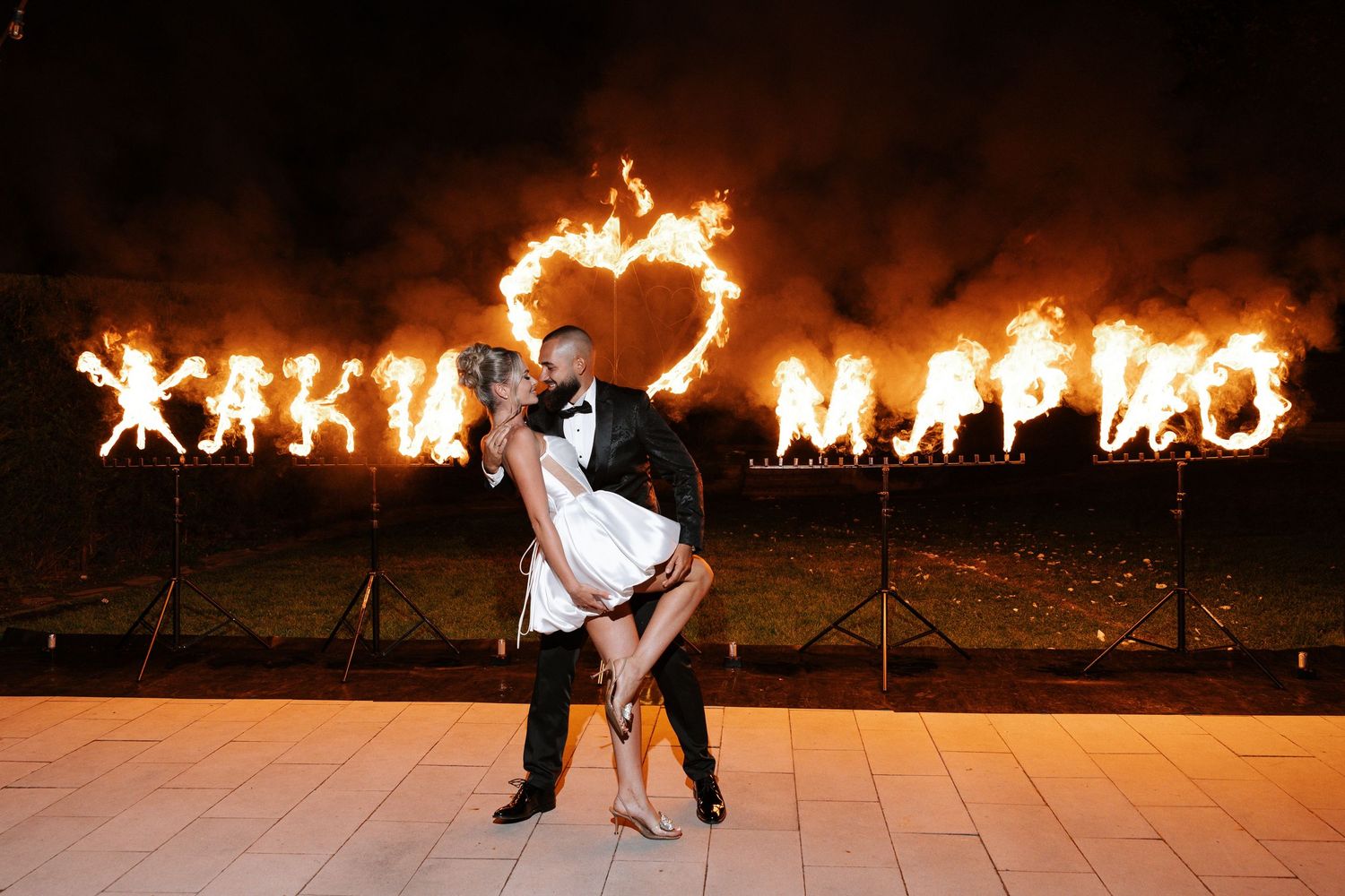 A couple dances in front of a flaming heart display at a romantic nighttime outdoor event.