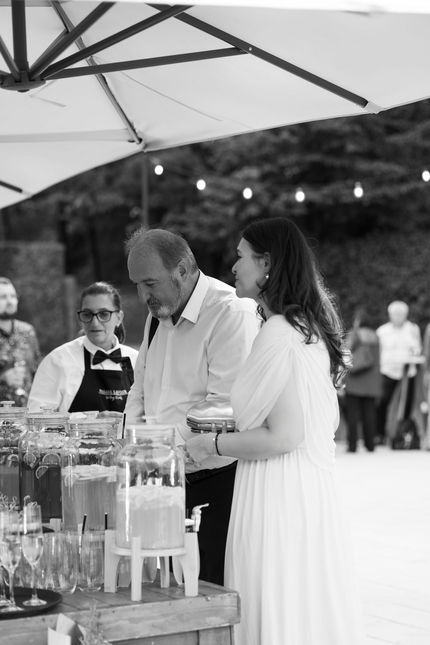 Wedding reception guests in white dresses gather around an outdoor beverage station with glass drink dispensers.