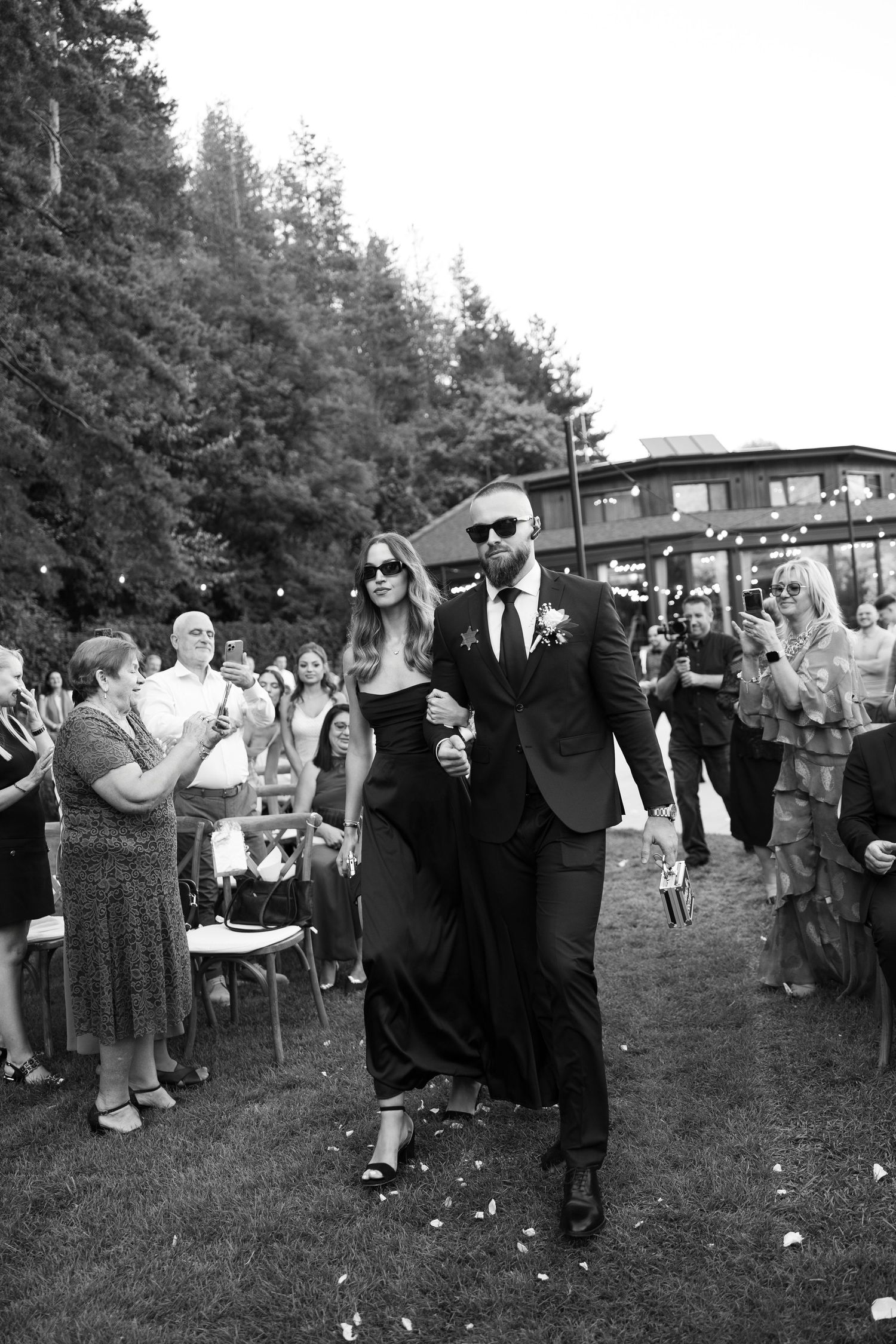 Black and white photo of wedding guests walking down an outdoor aisle while others watch and celebrate from the sides.