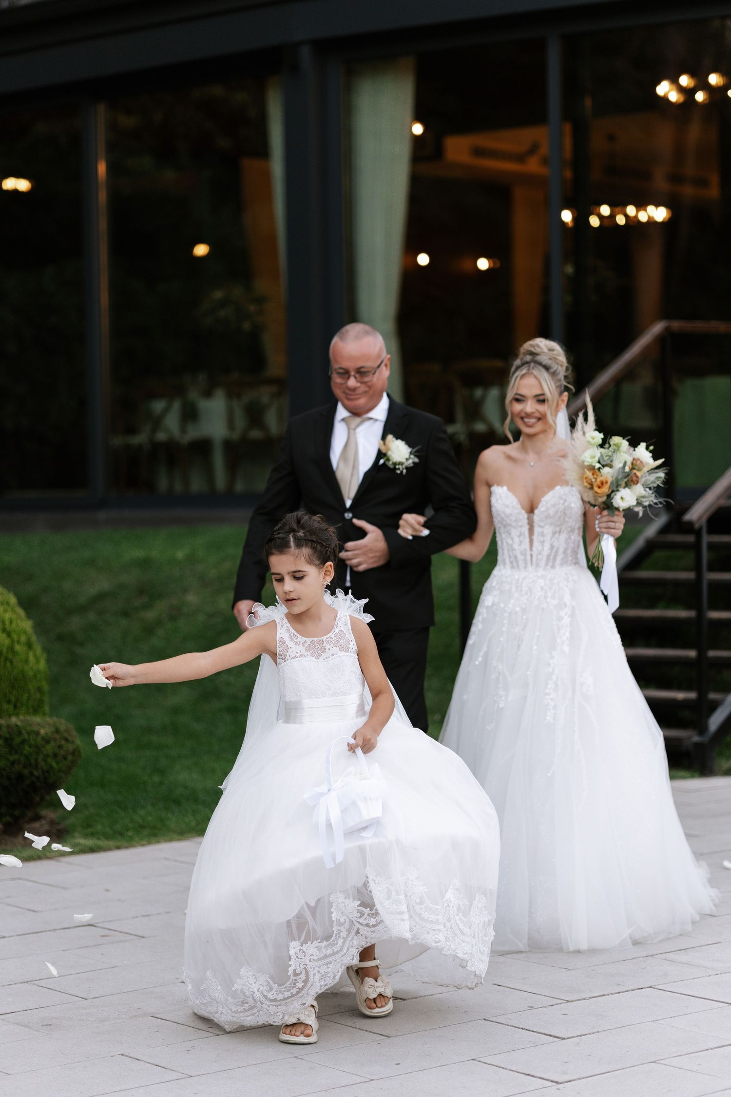 Two young flower girls in white dresses chase flower petals on a stone patio during a wedding celebration.