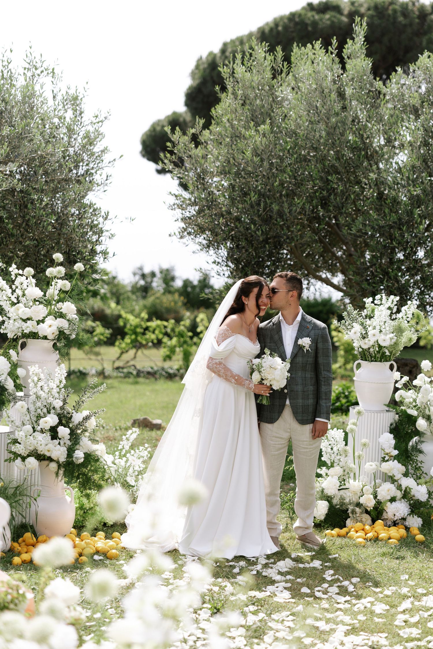 Romantic wedding couple poses in a garden setting with white floral arrangements and olive trees in the background.