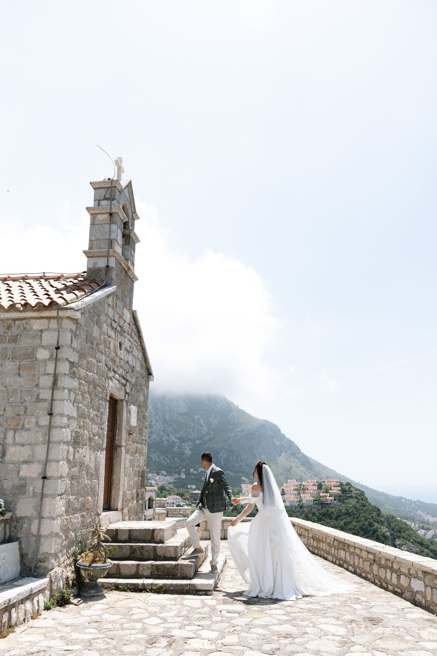 A couple stands on a stone terrace overlooking misty mountains at a historic fortress in Montenegro.
