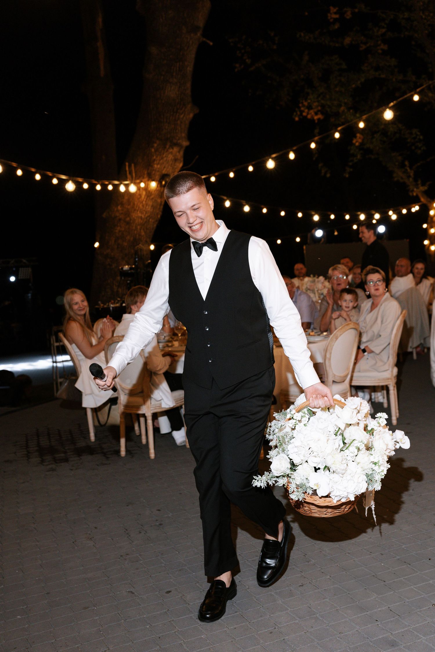 Wedding party member performing an energetic dance under string lights at an outdoor reception.