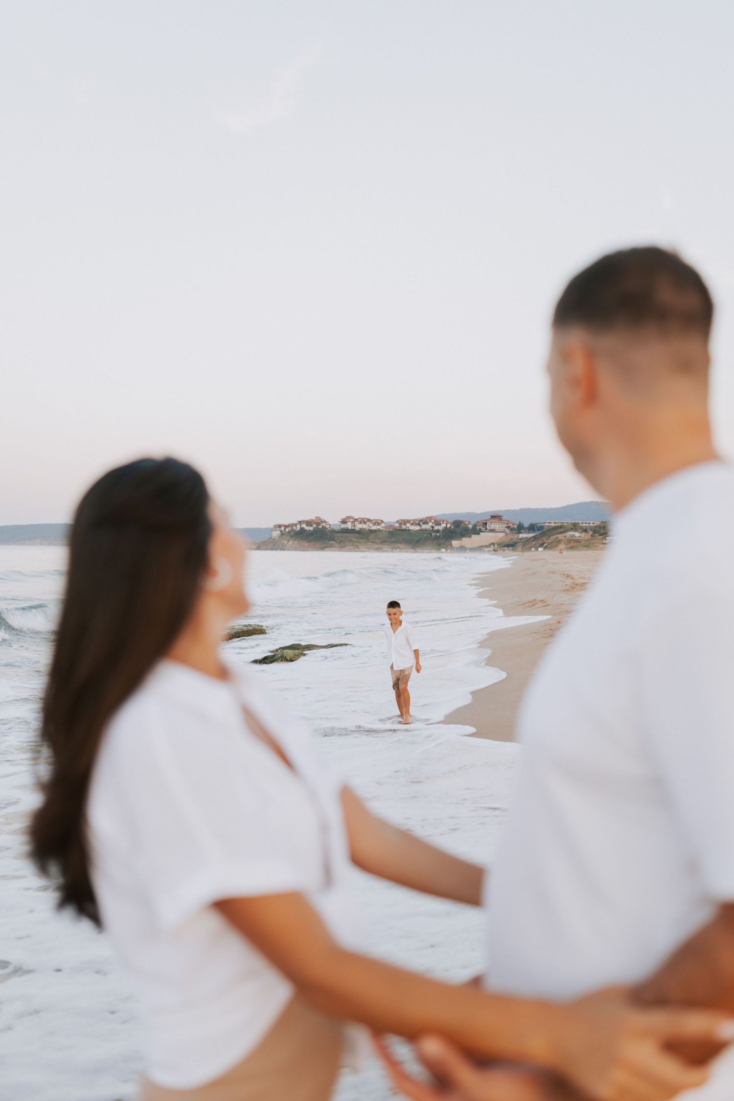 Three people in white clothing pose for photos on a scenic beach with coastal cliffs in the distance.