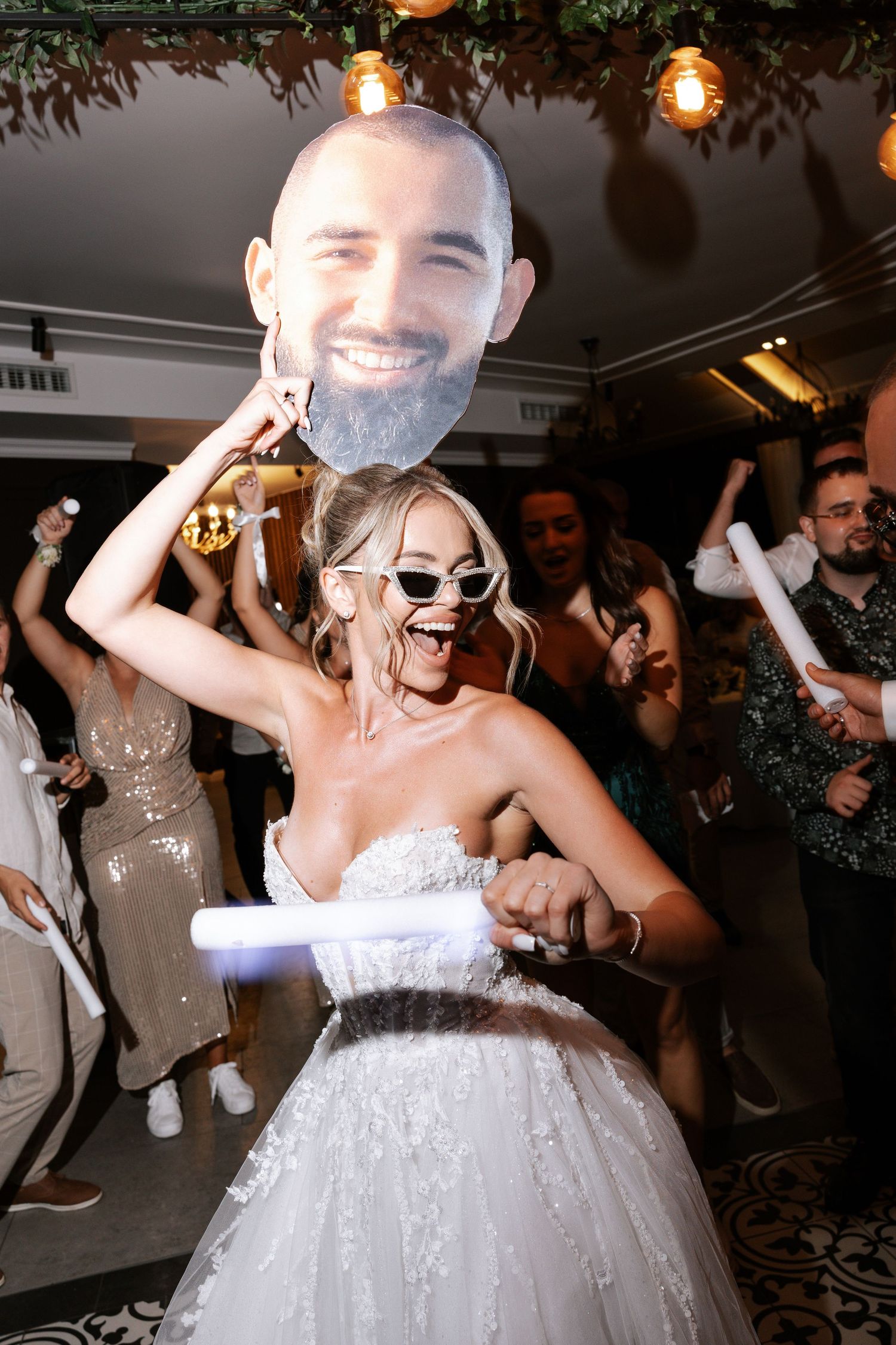 A bride in a white wedding dress and sunglasses dances at a reception while holding a decorative prop under hanging lights.