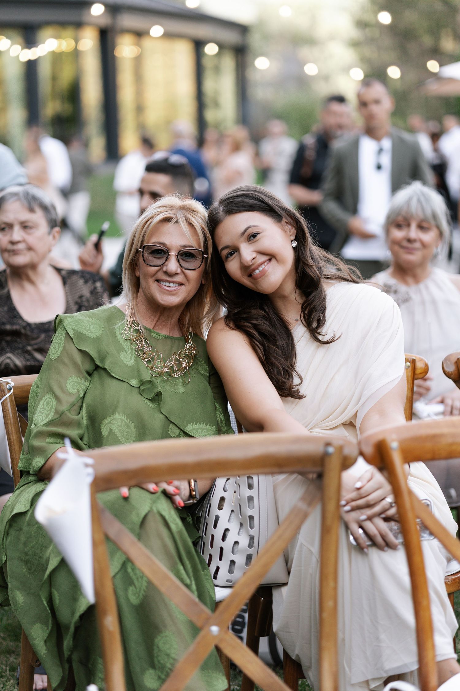 Two people in green and white outfits share a candid moment at an outdoor dining event with wooden chairs.