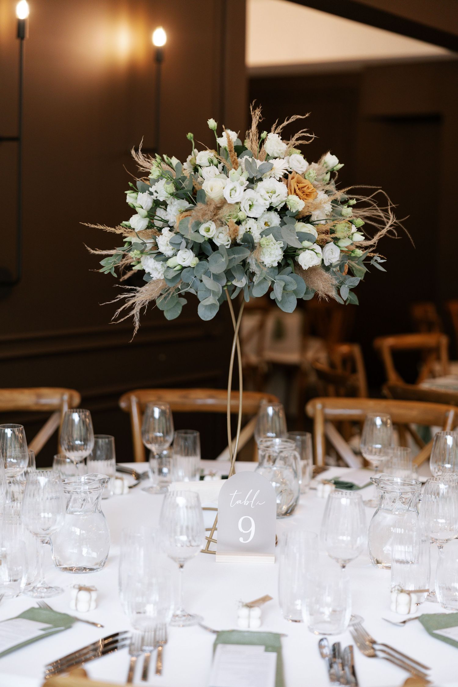 Elegant wedding centerpiece with white flowers and eucalyptus on tall stand above formal place settings at reception table.