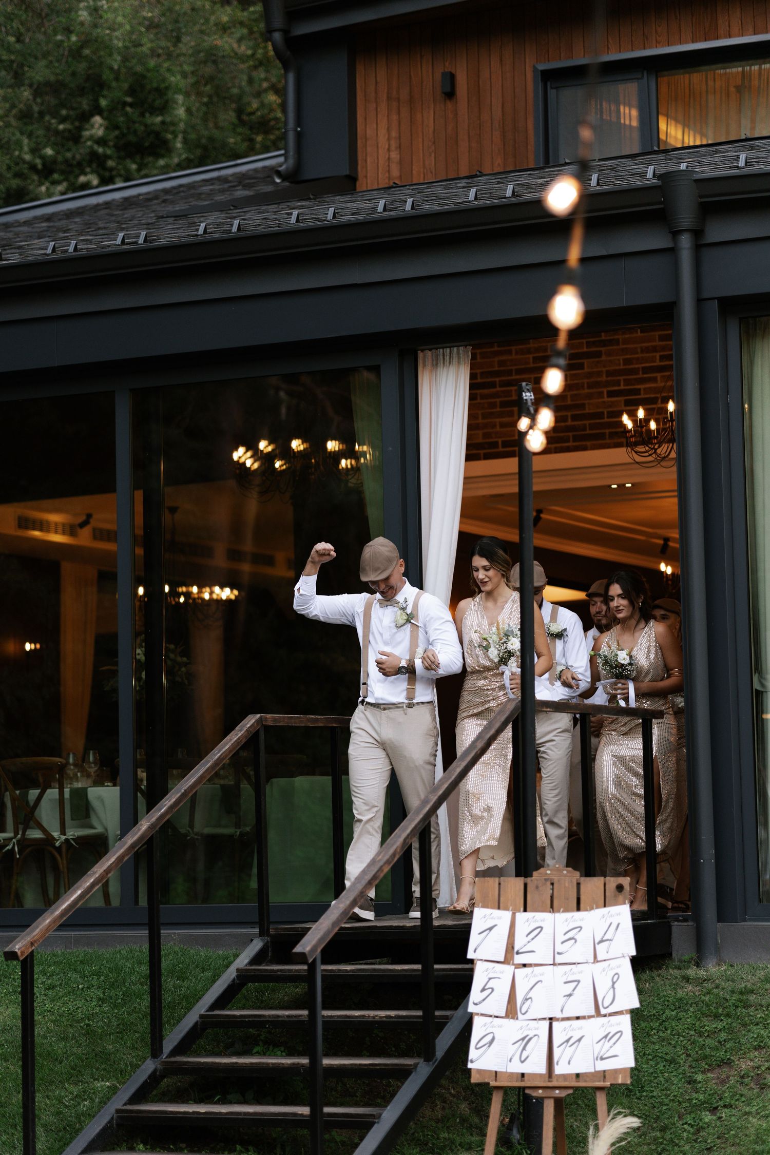 Wedding party poses with cardboard signs in sequence outside rustic wooden venue with string lights at dusk.