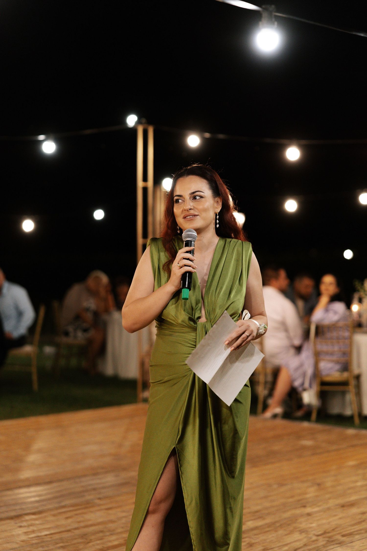 Someone in an olive green dress speaks into a microphone on a wooden dance floor under string lights at an outdoor event.