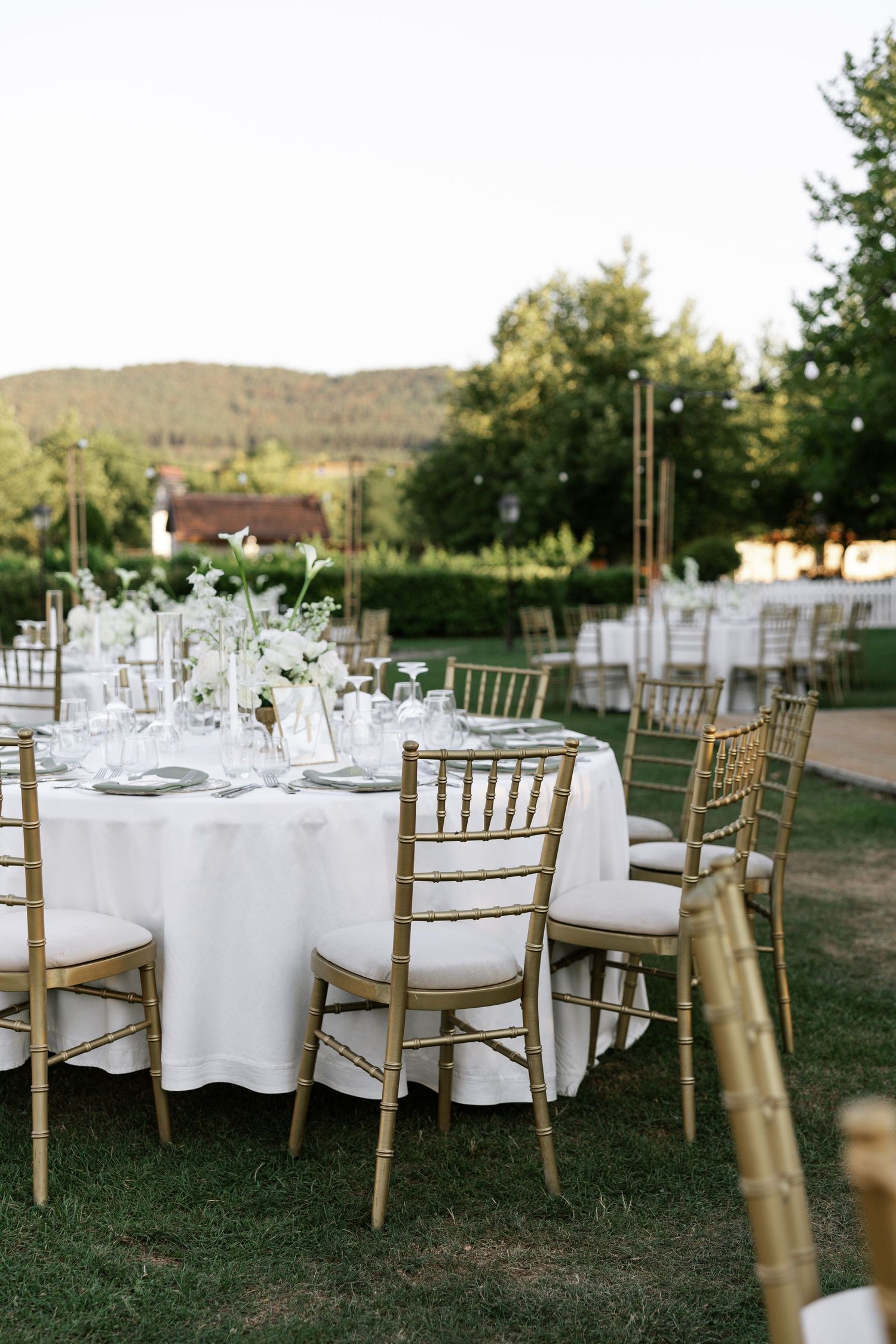Elegant outdoor wedding reception setup with round tables, white linens, and gold chiavari chairs on a grassy lawn.