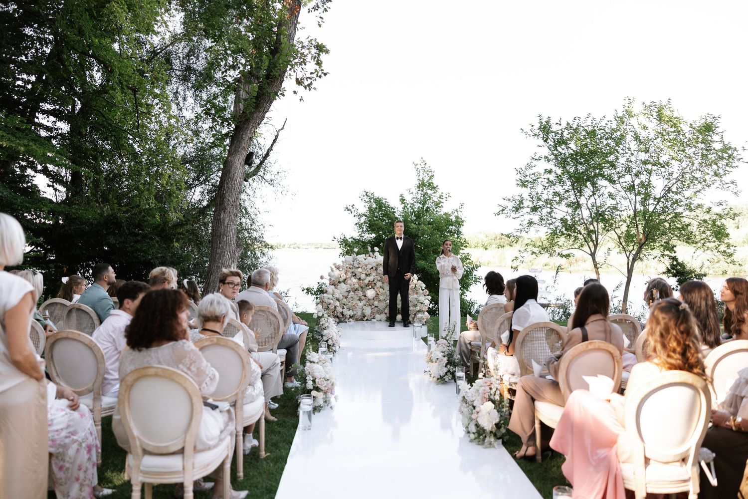 Outdoor wedding ceremony setup with white chairs and aisle runner surrounded by trees.