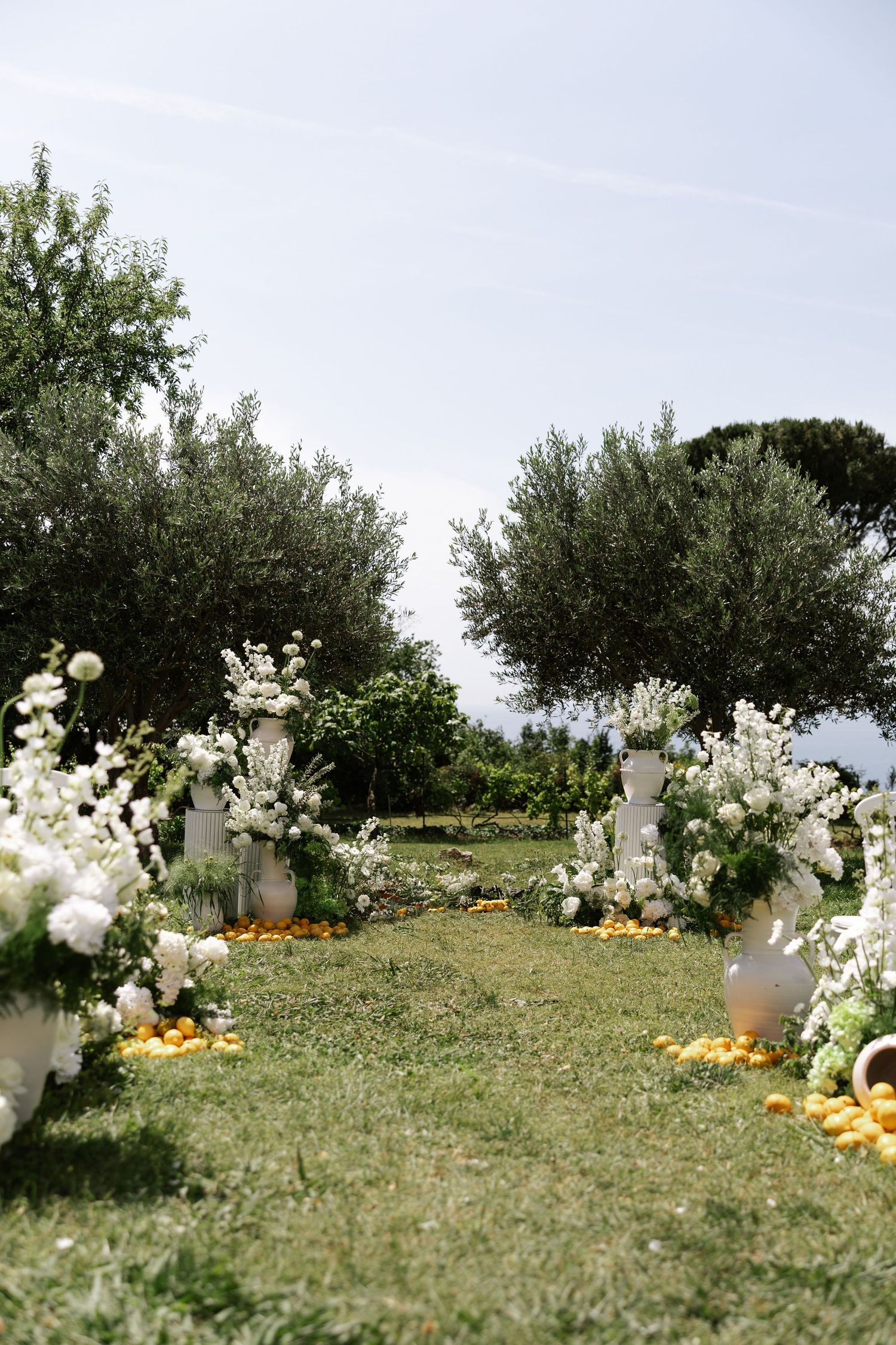 White floral arrangements and pumpkins line a grassy outdoor path beneath olive trees creating an elegant garden setting.