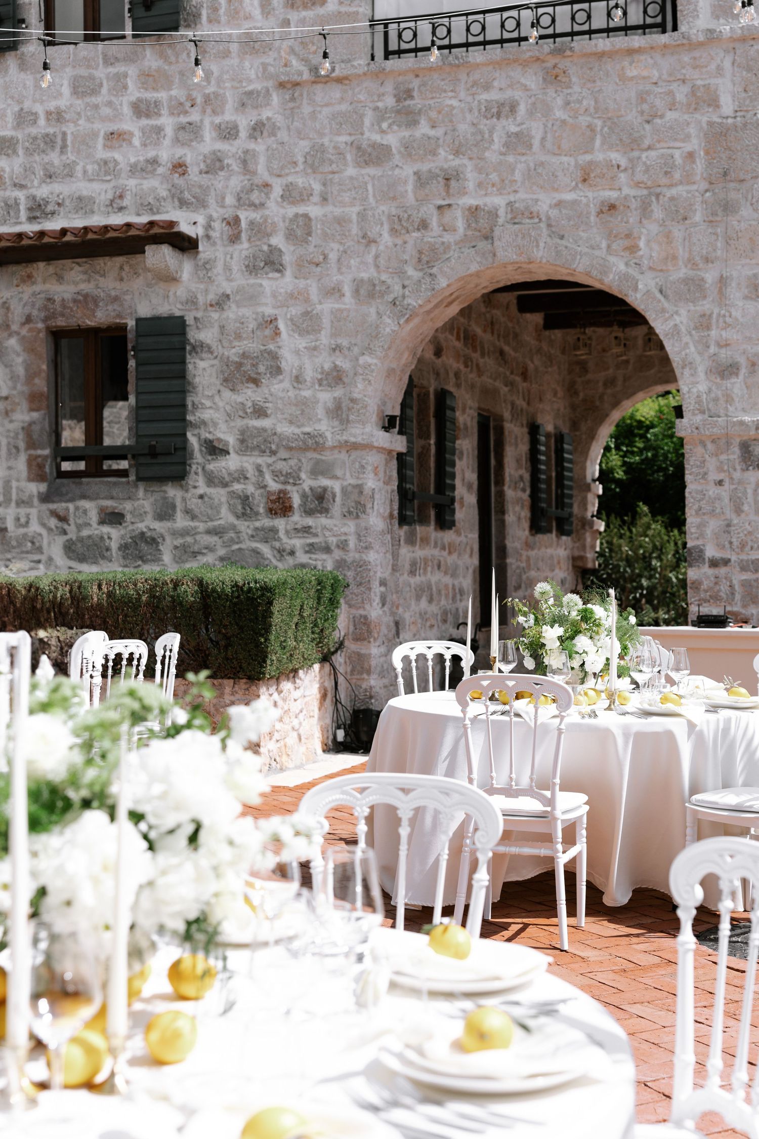 Elegant outdoor wedding reception setup with white chairs and tables under stone archways of a rustic Mediterranean venue.