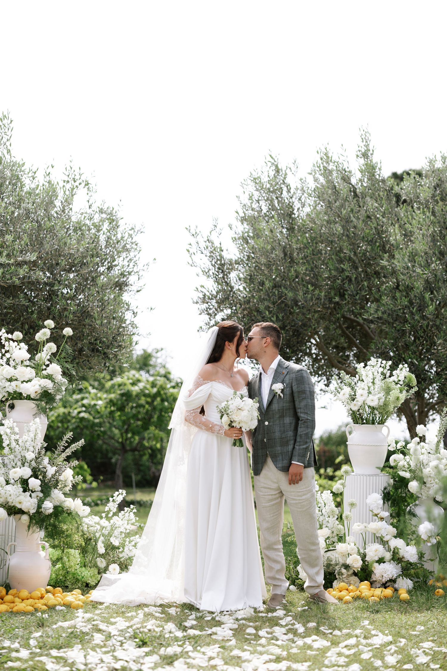 A romantic outdoor wedding ceremony scene with white floral arrangements and olive trees in a Mediterranean setting.