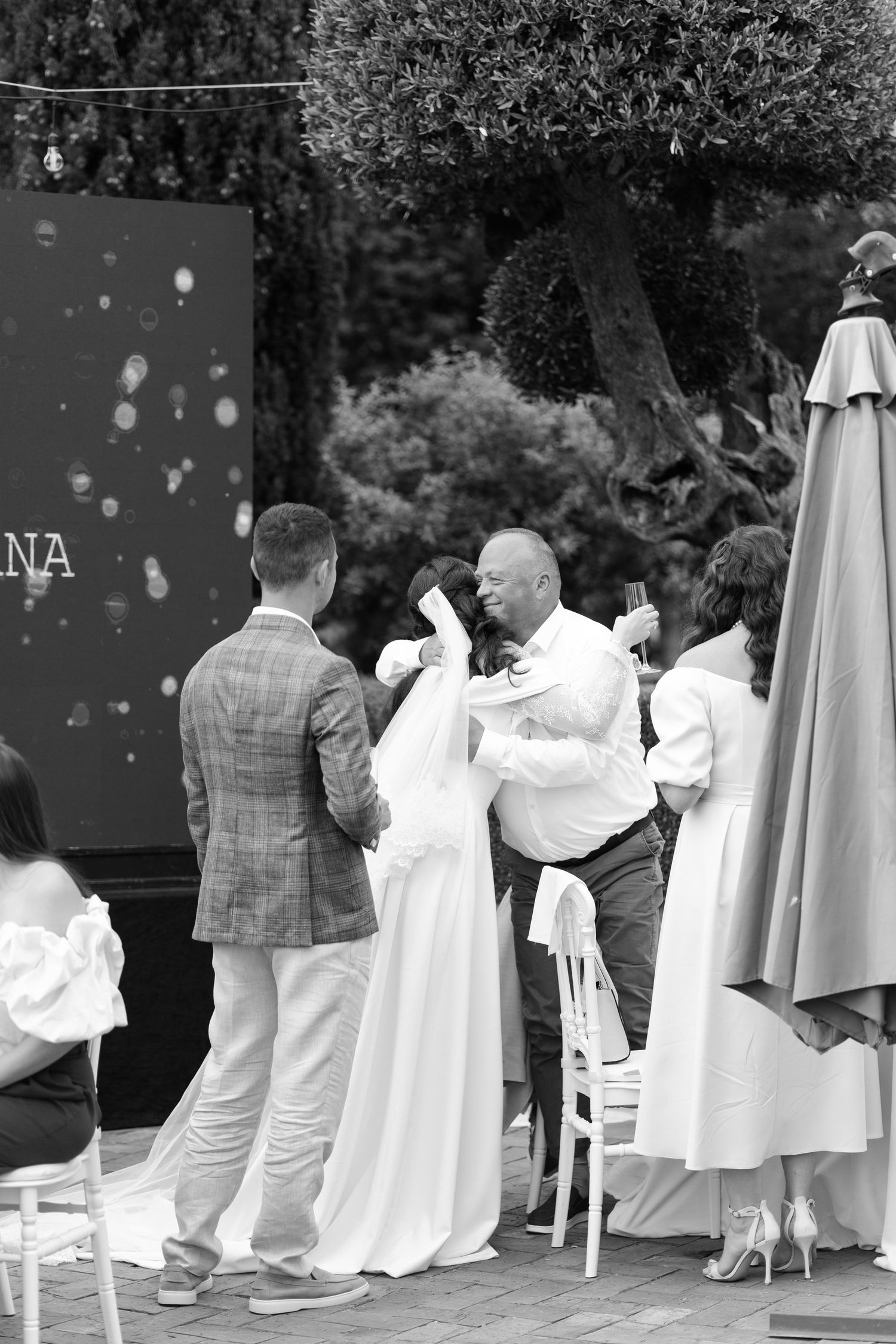 A black and white candid photograph of a wedding ceremony taking place outdoors with guests and umbrellas visible.