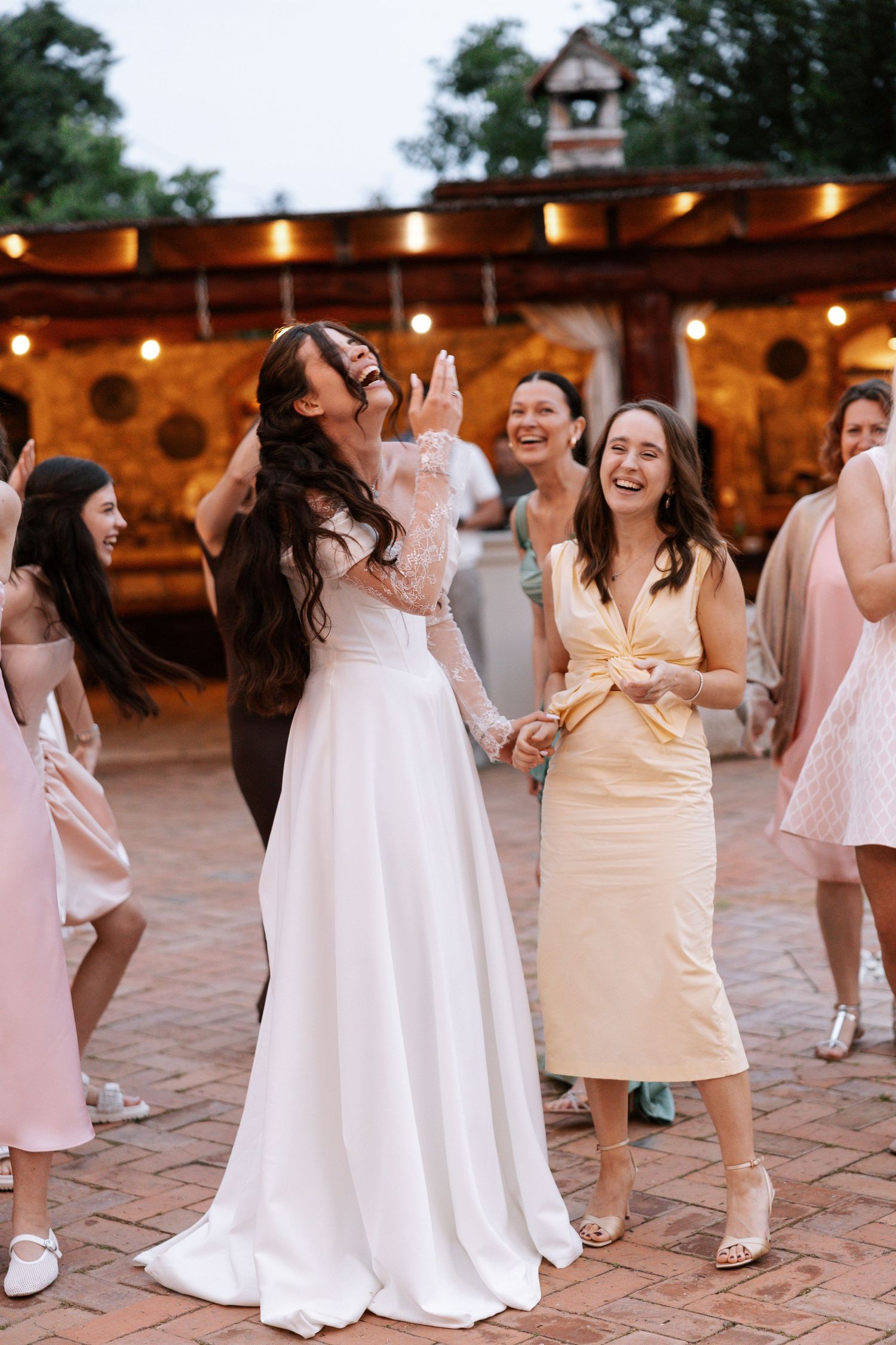 Wedding party dances and laughs together on outdoor brick patio with string lights at evening reception.