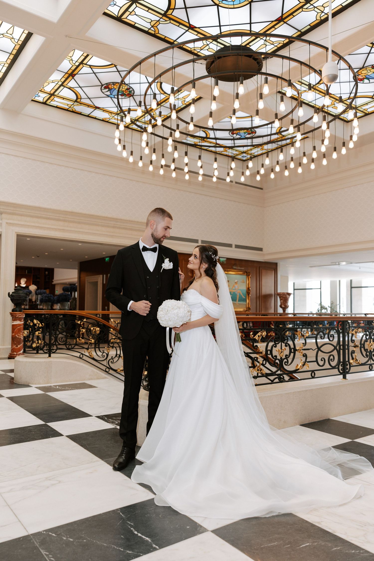 Wedding couple stands beneath stunning circular chandelier on black and white marble floor.