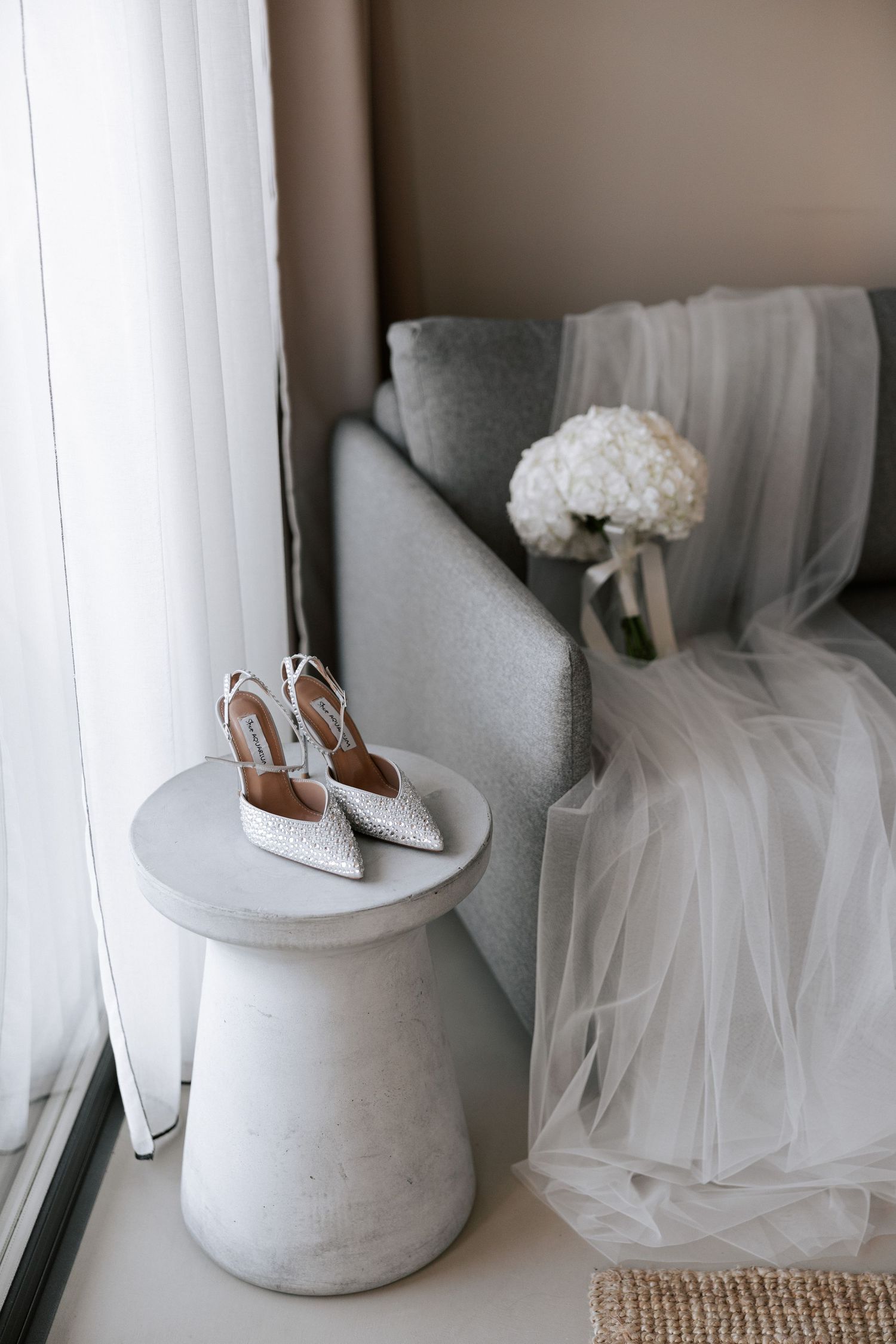 Wedding shoes and white bouquet displayed on concrete side table near window.