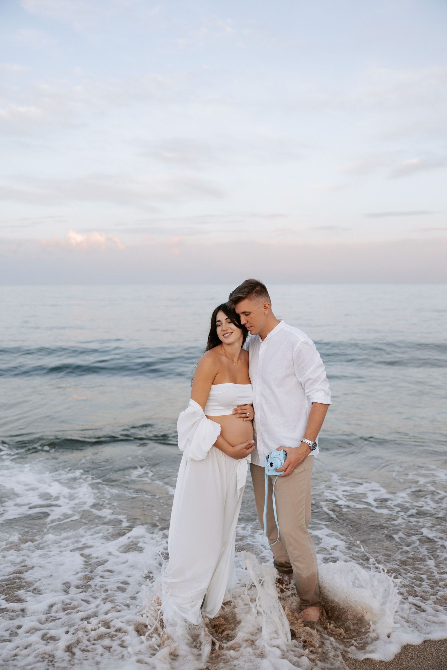 A tender moment between expecting parents standing in ocean waves at sunset.