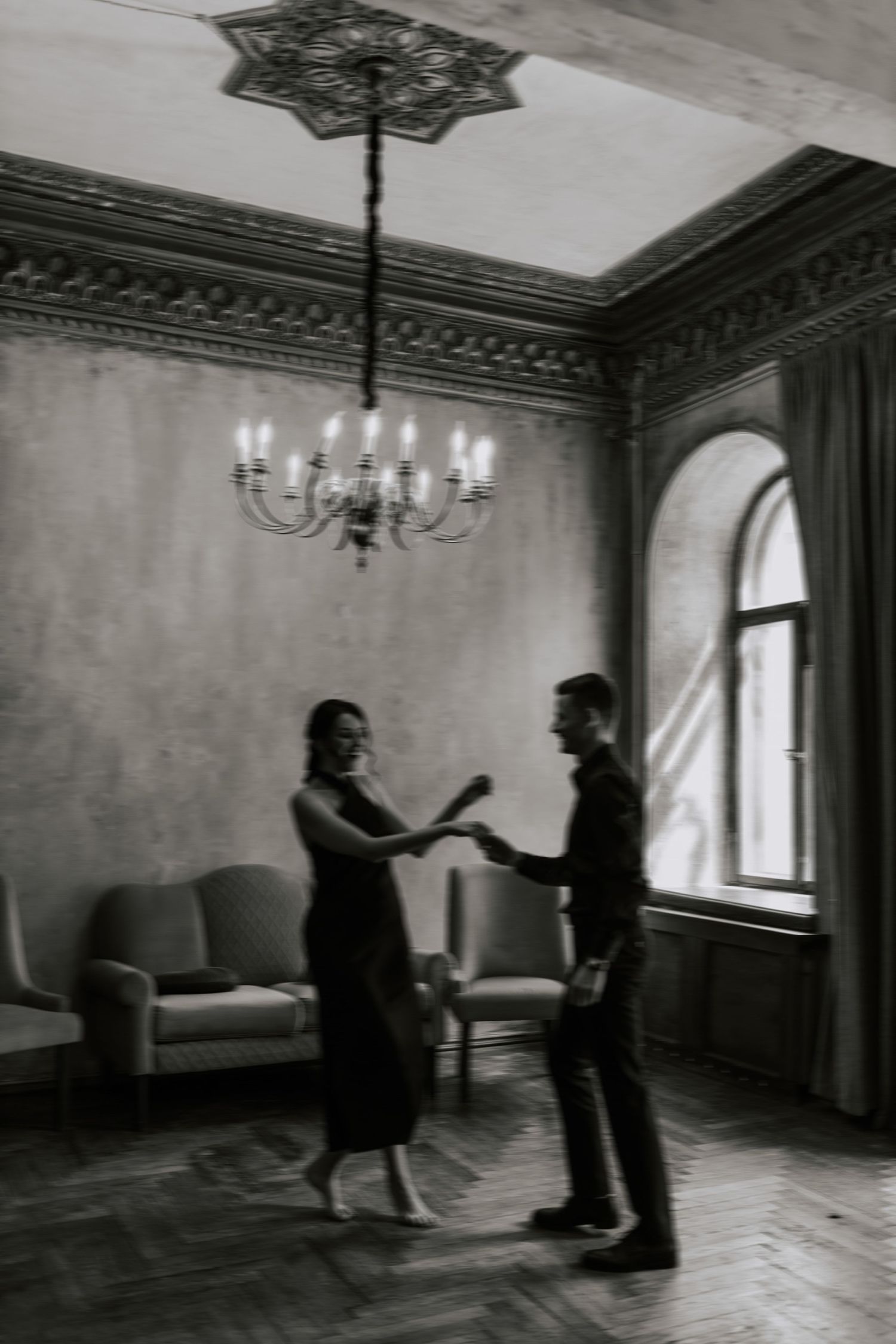Two people dance together in an elegant room with chandelier and arched windows in black and white.