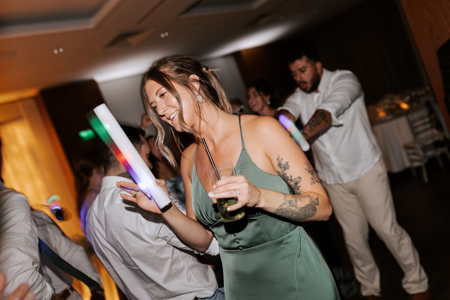 Woman in sage green dress dances while holding a light-up party favor at wedding reception.