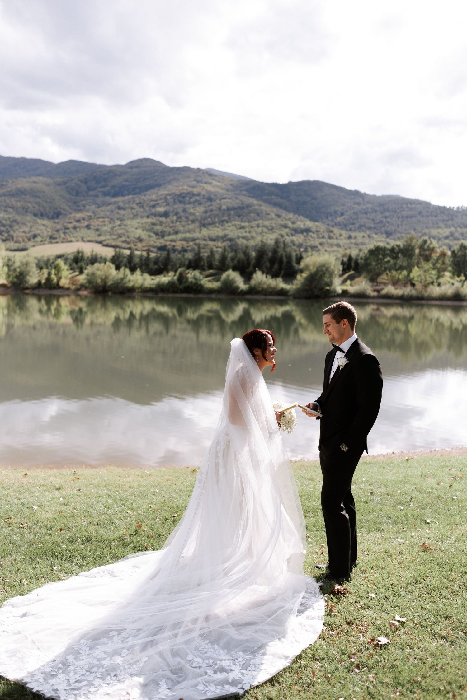 A bride's dramatic cathedral veil trails behind her as the couple stands by a mountain lake.