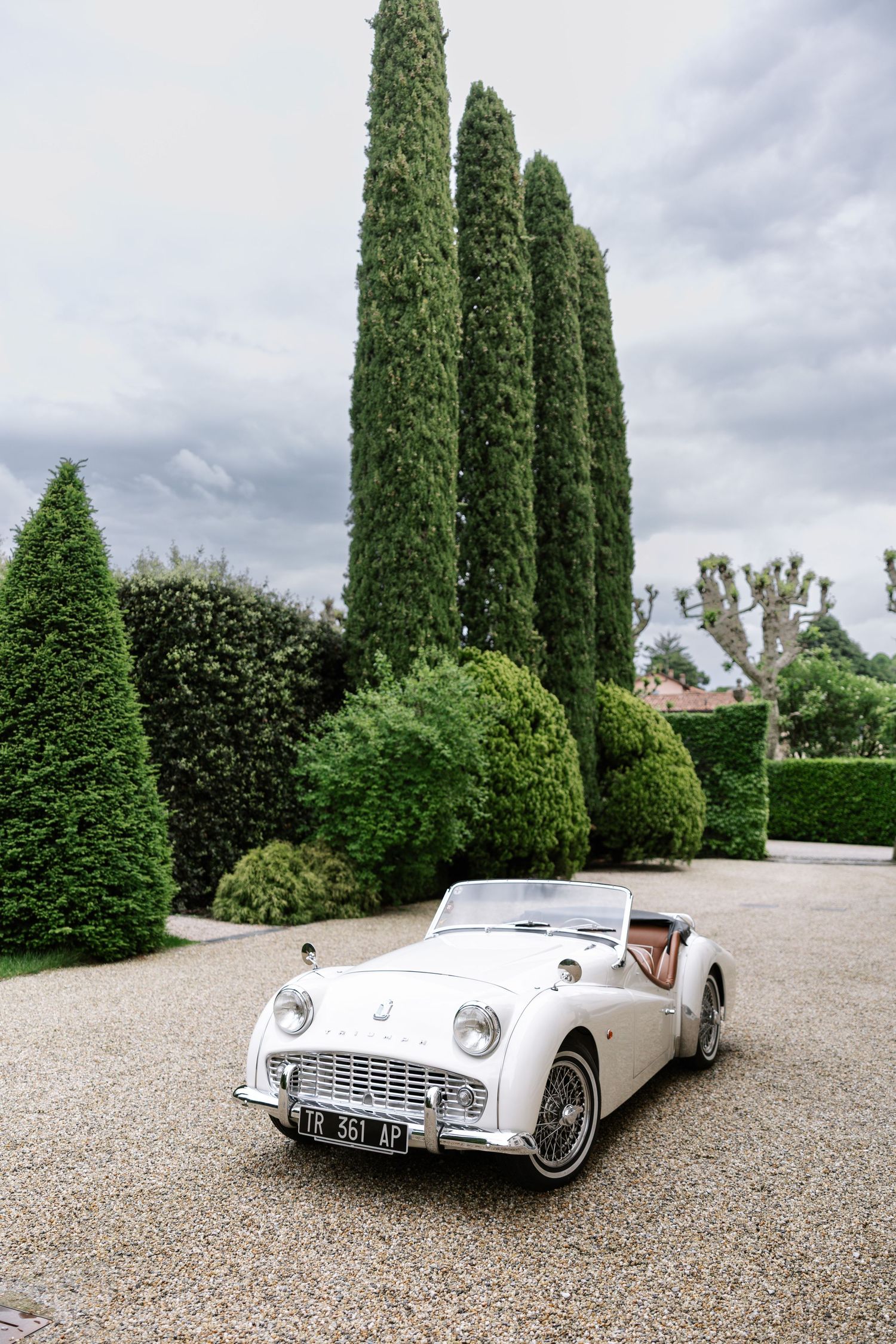 Classic white convertible sports car parked on gravel driveway with tall cypress trees in the background.