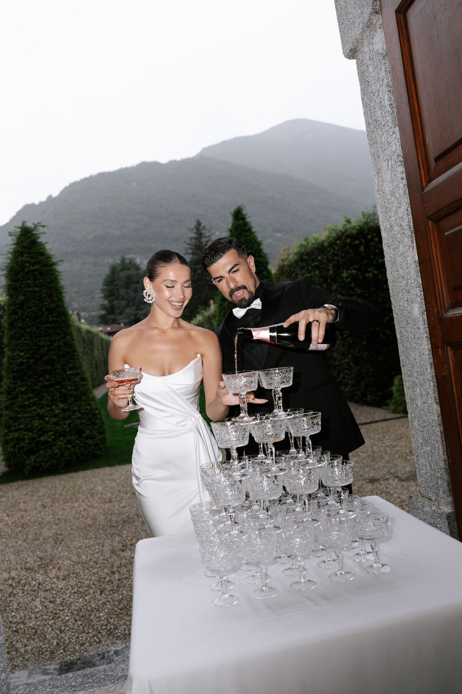 Wedding couple pours champagne into a tower of crystal glasses stacked in a pyramid formation at an outdoor venue.