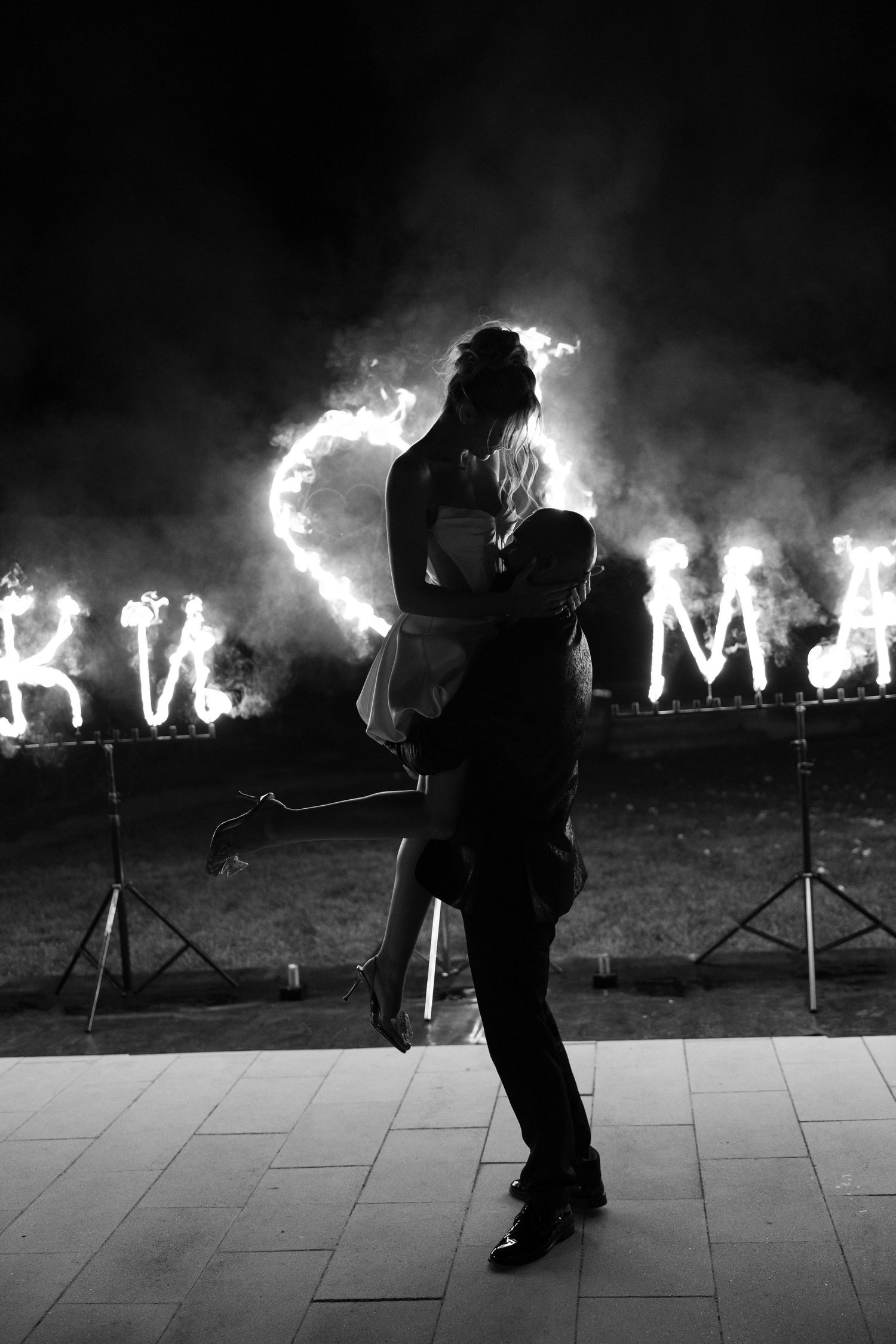 Silhouettes dance against a dramatic backdrop of burning flames arranged in a line during a nighttime performance.