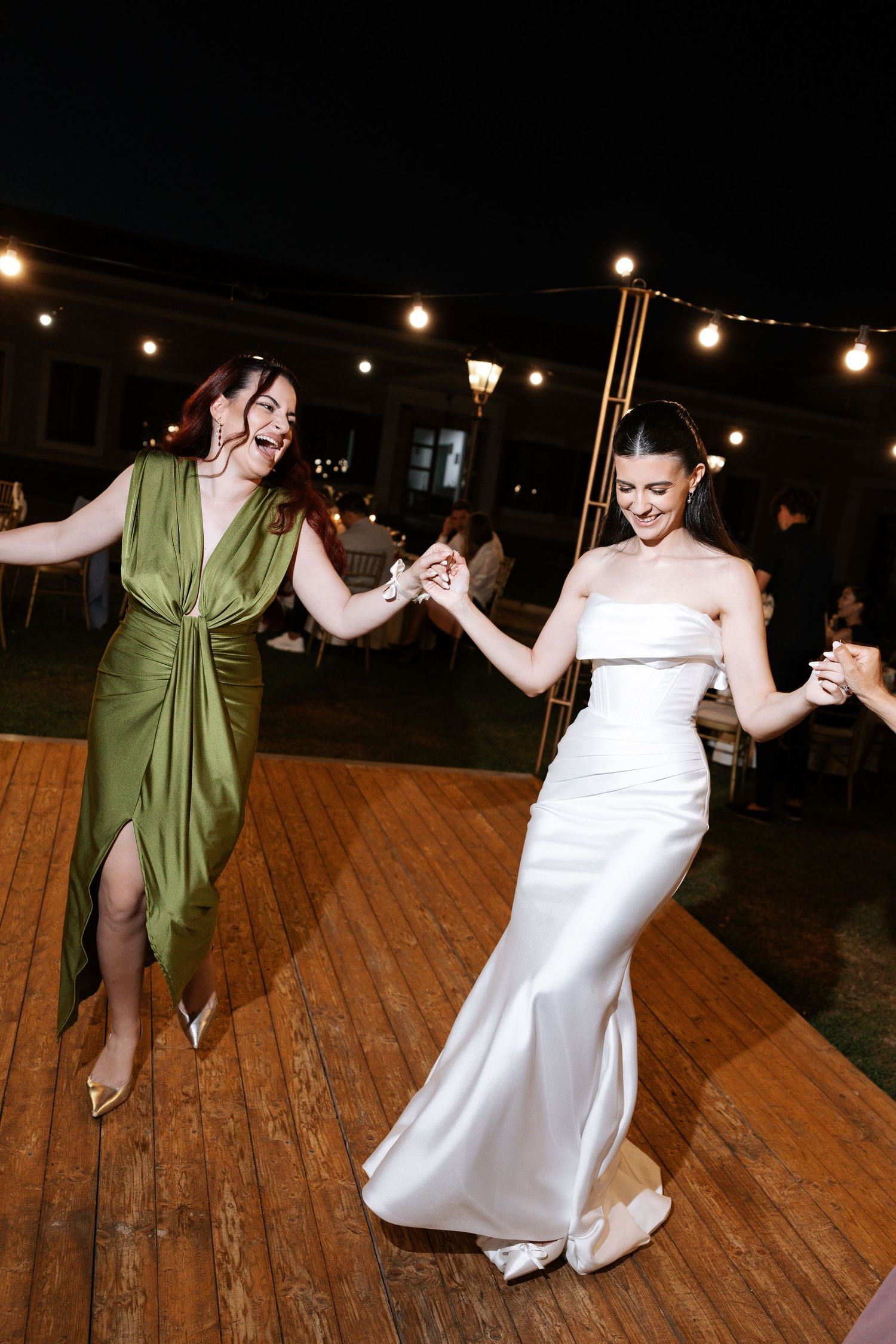 Two people in formal attire dancing together on a wooden deck under string lights at night.