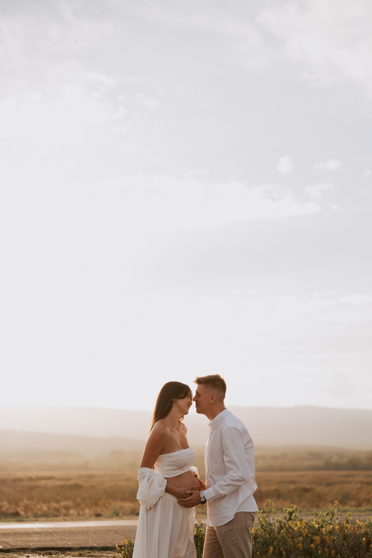 Romantic maternity photo of couple embracing against misty landscape at sunset.
