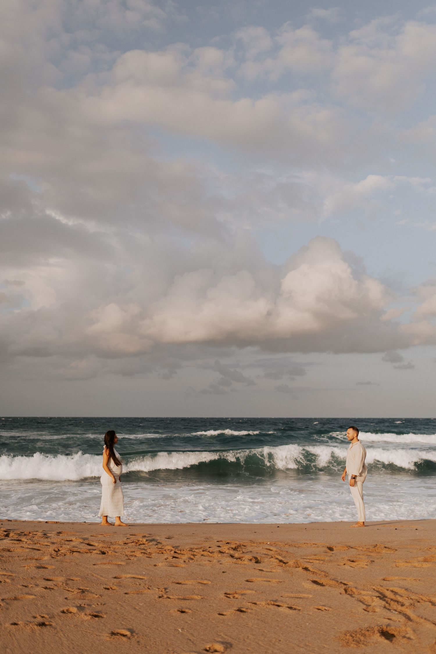 Two people in white outfits standing apart on a beach with turbulent ocean waves.