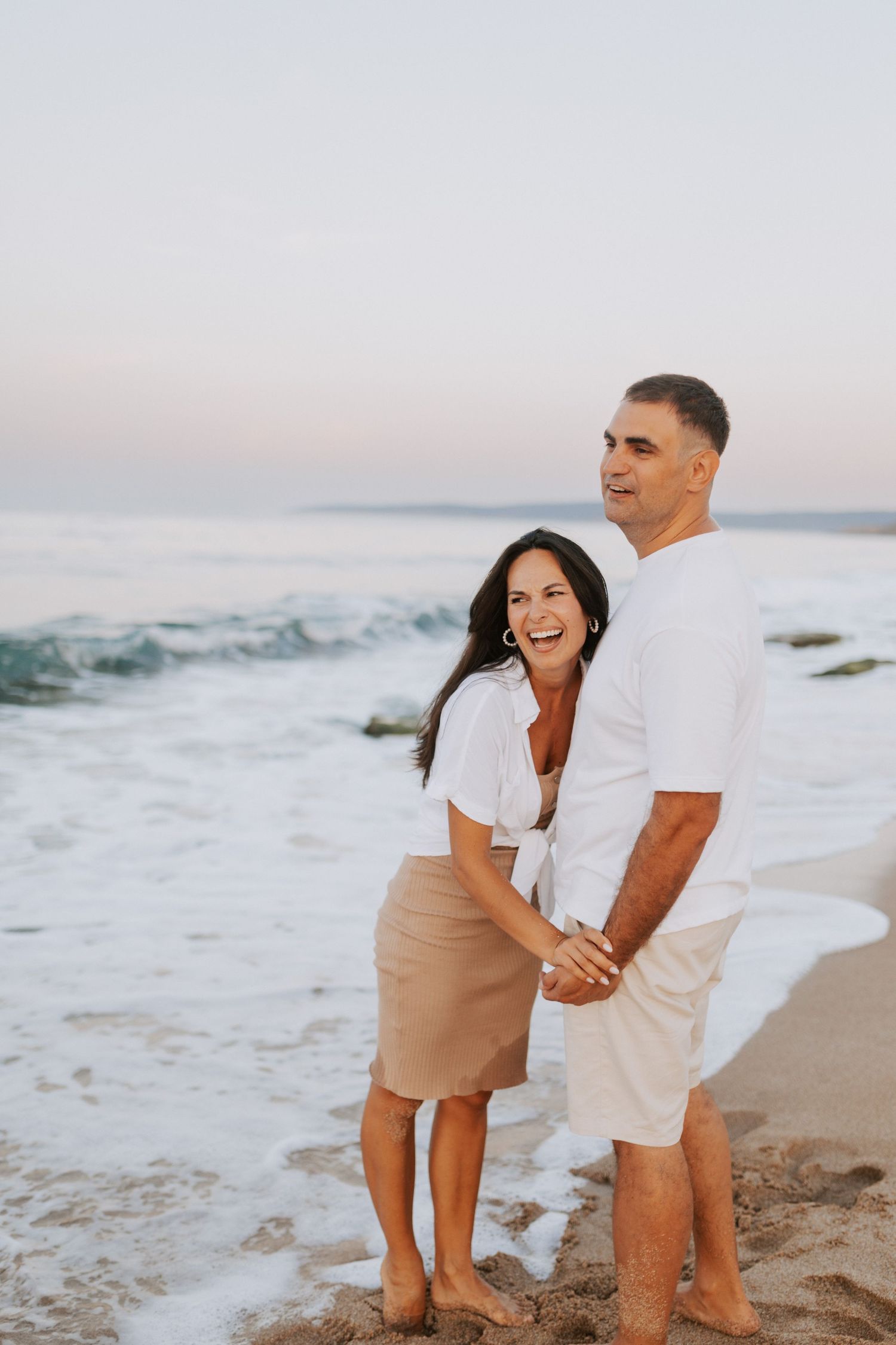 A couple shares a joyful moment together on a beach at sunset with crashing waves in the background.