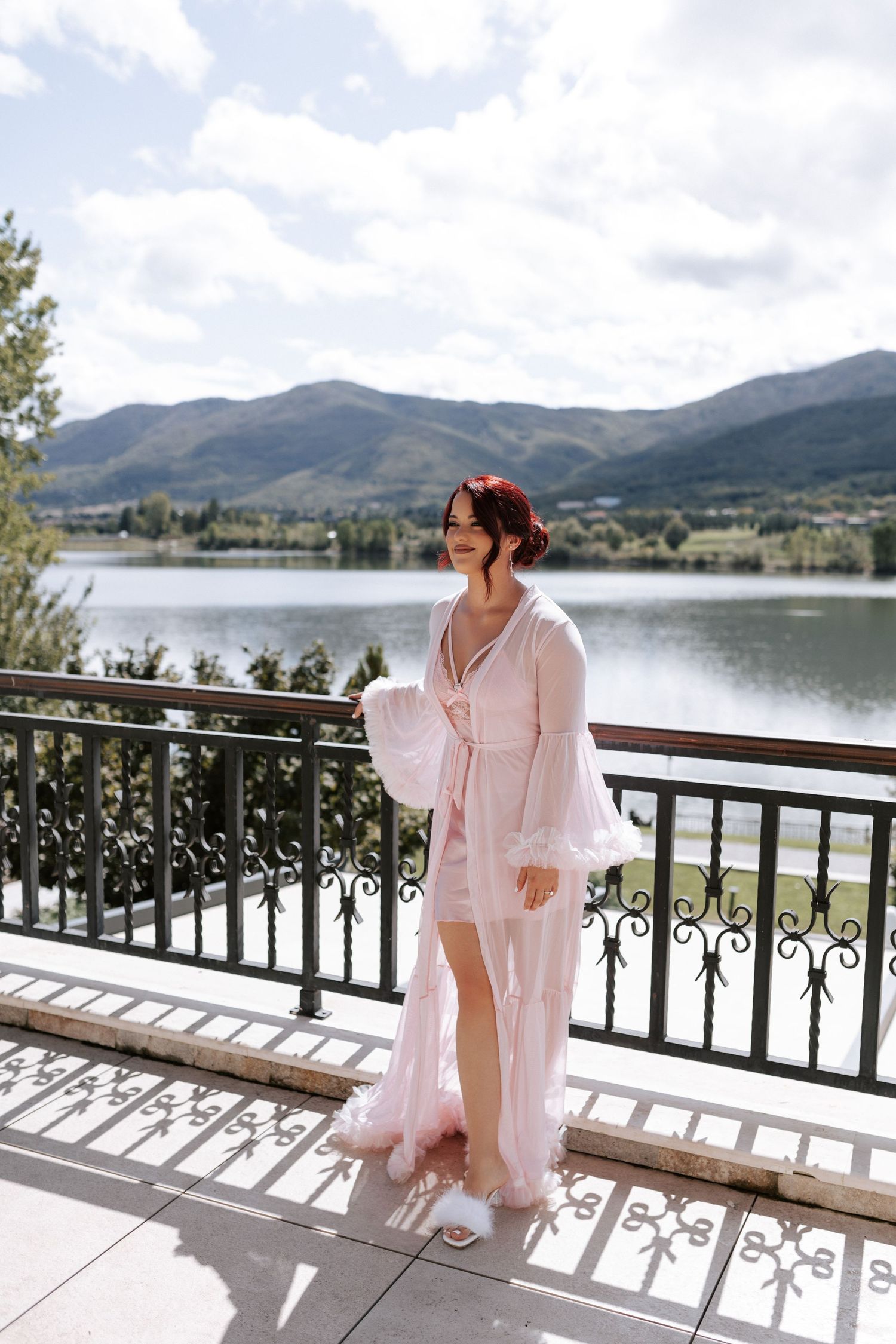 Person in pink flowing robe standing on balcony with mountain lake view.