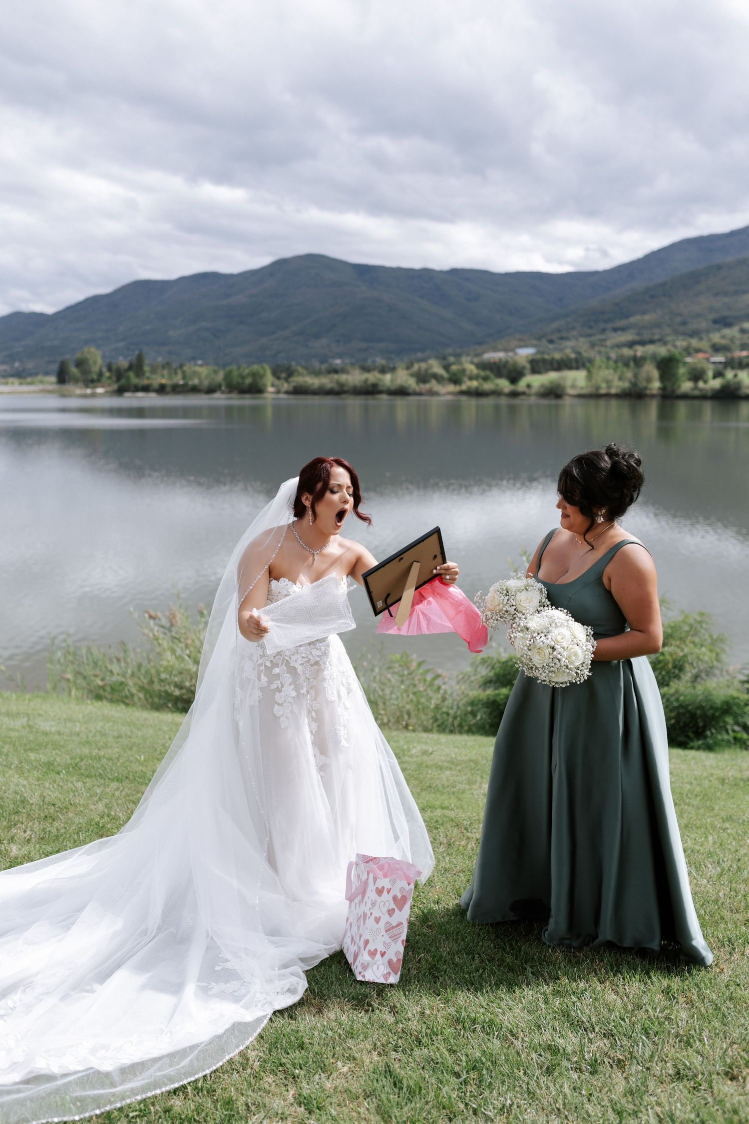 Bride in lace gown views special gift with bridesmaid by mountain lake setting.