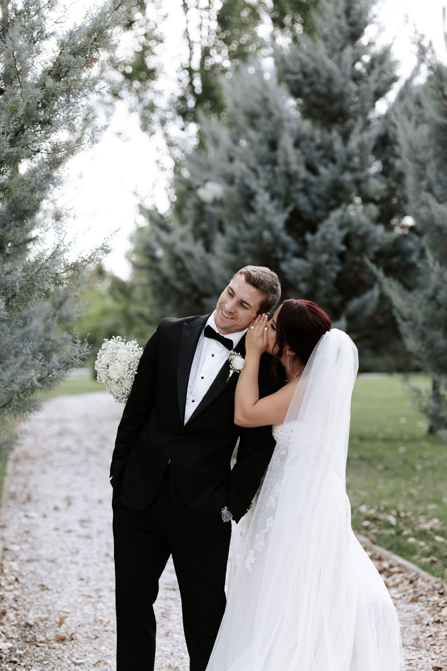 Intimate wedding moment captured between couple along evergreen-lined path.